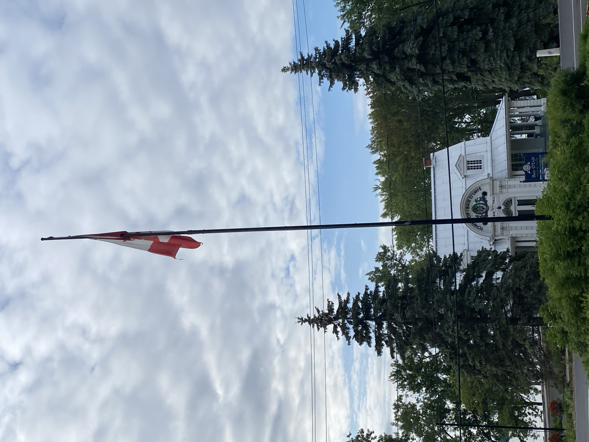 This media file captures a scene featuring a Canadian flag flying prominently against a cloudy sky. The flag is positioned on a tall flagpole to the left of the frame, its red and white design clearly visible. In the background, a white building with columns and a pitched roof stands behind a line of tall evergreen trees and deciduous trees. The building appears to be a public institution, as indicated by the text visible on a blue sign and the building itself. The sign reads "Bar-Wcff". The building also has text that reads "OF-BAIE D'URFÉ" and "TOWN 1917 HALL". The scene is set outdoors, with overhead power lines crossing the sky, suggesting an urban or suburban environment. The weather appears to be overcast but with patches of blue sky peeking through, indicating a partly cloudy day. The lighting suggests it is daytime, likely morning or afternoon. The overall impression is one of a civic location, possibly a town hall, in Baie-D'Urfé, Canada.