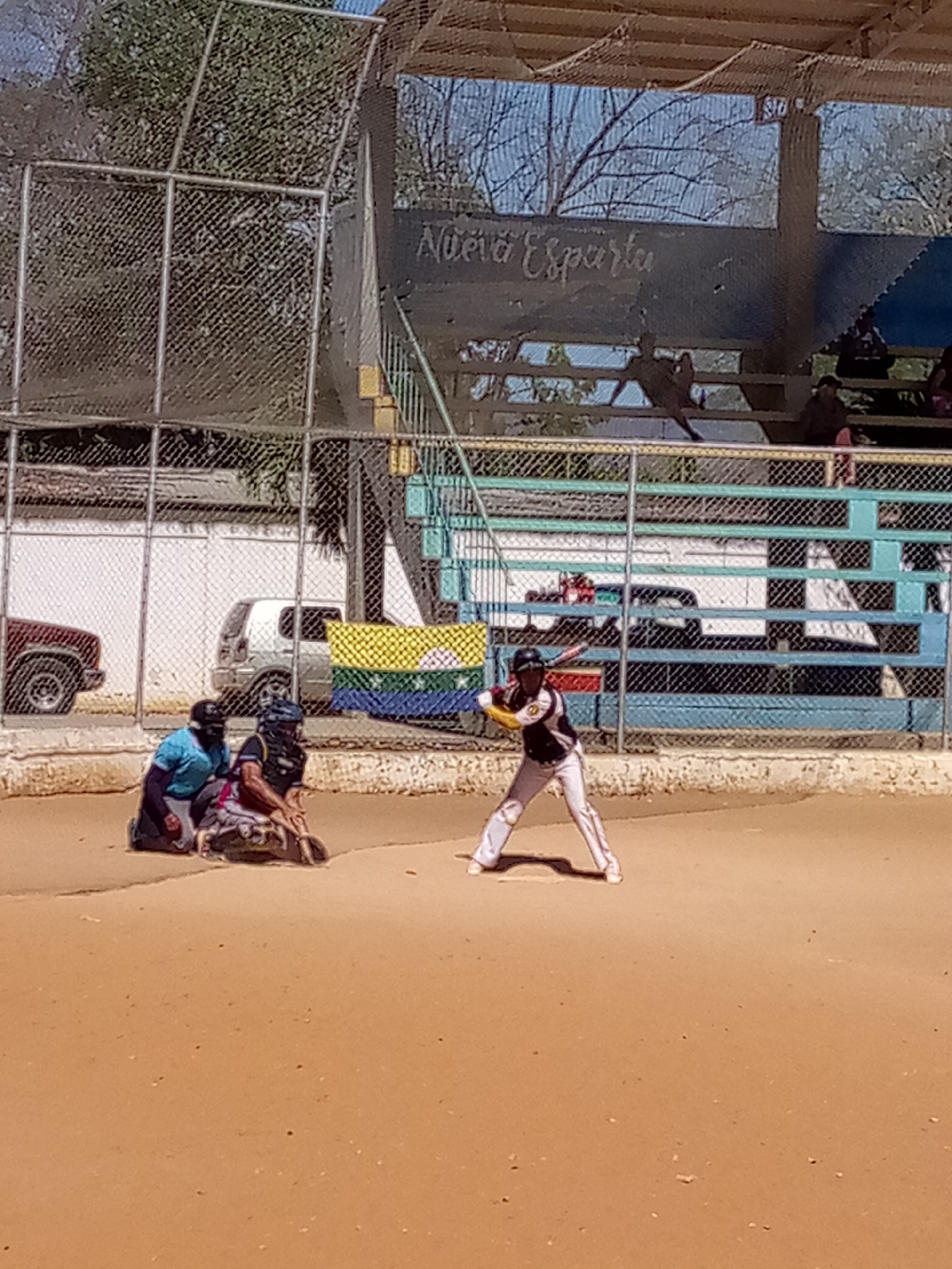A baseball game is in progress at a baseball field in Unknown, Unknown. A batter in white pants and a black shirt stands at home plate, holding a yellow bat. A catcher in a black helmet and padding is crouched behind the batter. Next to the catcher is another person in a blue shirt and a black helmet. A wire fence surrounds the field. Behind the fence are cars and a building with a Venezuelan flag hanging on it. In the background is a small stadium with the words "Nueva Esparta" written on it. There are a few people sitting in the stadium. The baseball field is made of dirt. The sky is visible above the stadium.