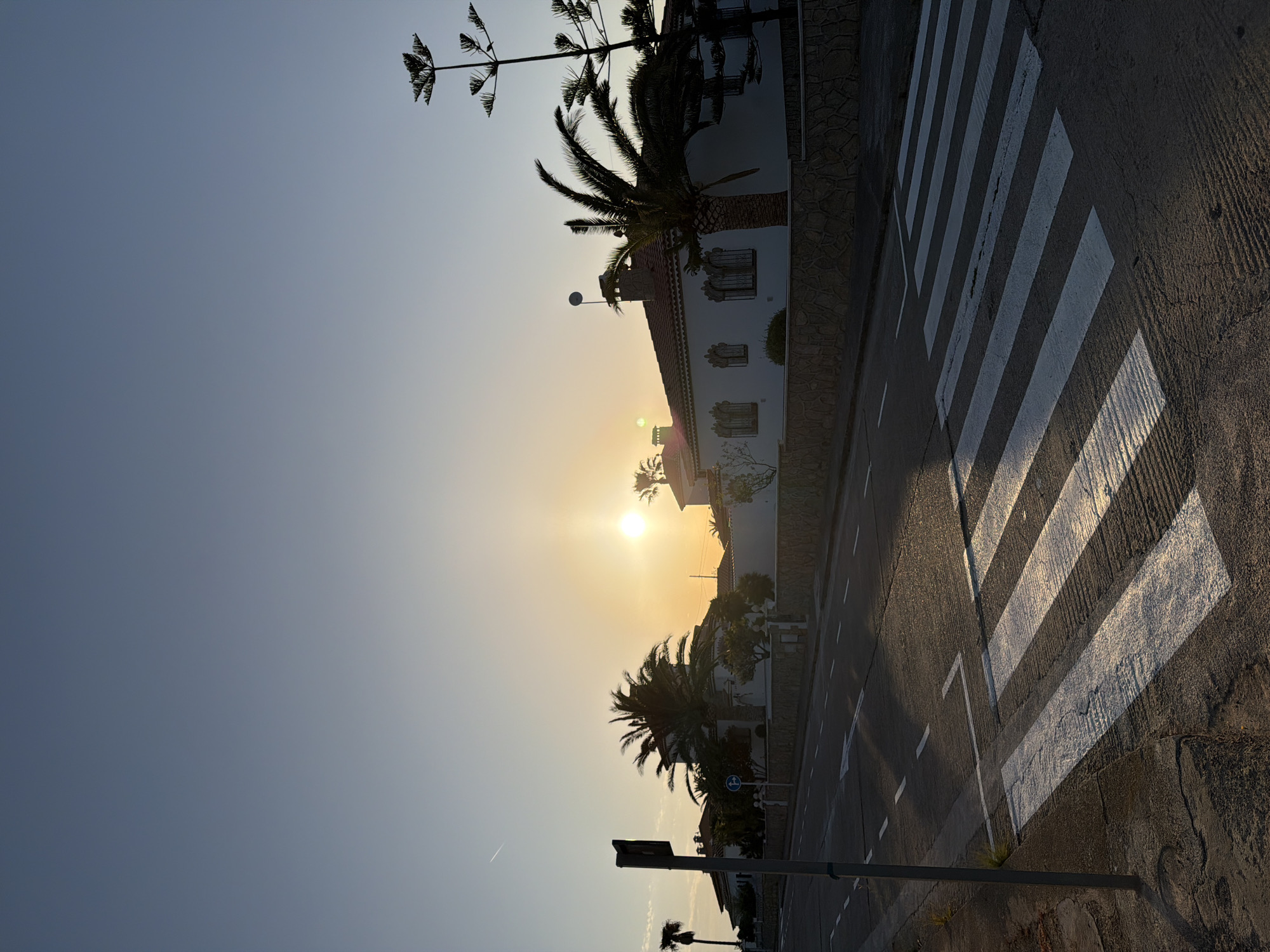 The image depicts a street scene in Mont-roig del Camp, Spain, during sunset. The sun is positioned low in the sky, causing a bright glare. There is a zebra crossing running across the road. Buildings are visible, including white structures with tiled roofs and stone walls. Palm trees and other vegetation add a tropical feel. A street light and road signs are present. The road is asphalt, showing wear and tear.
