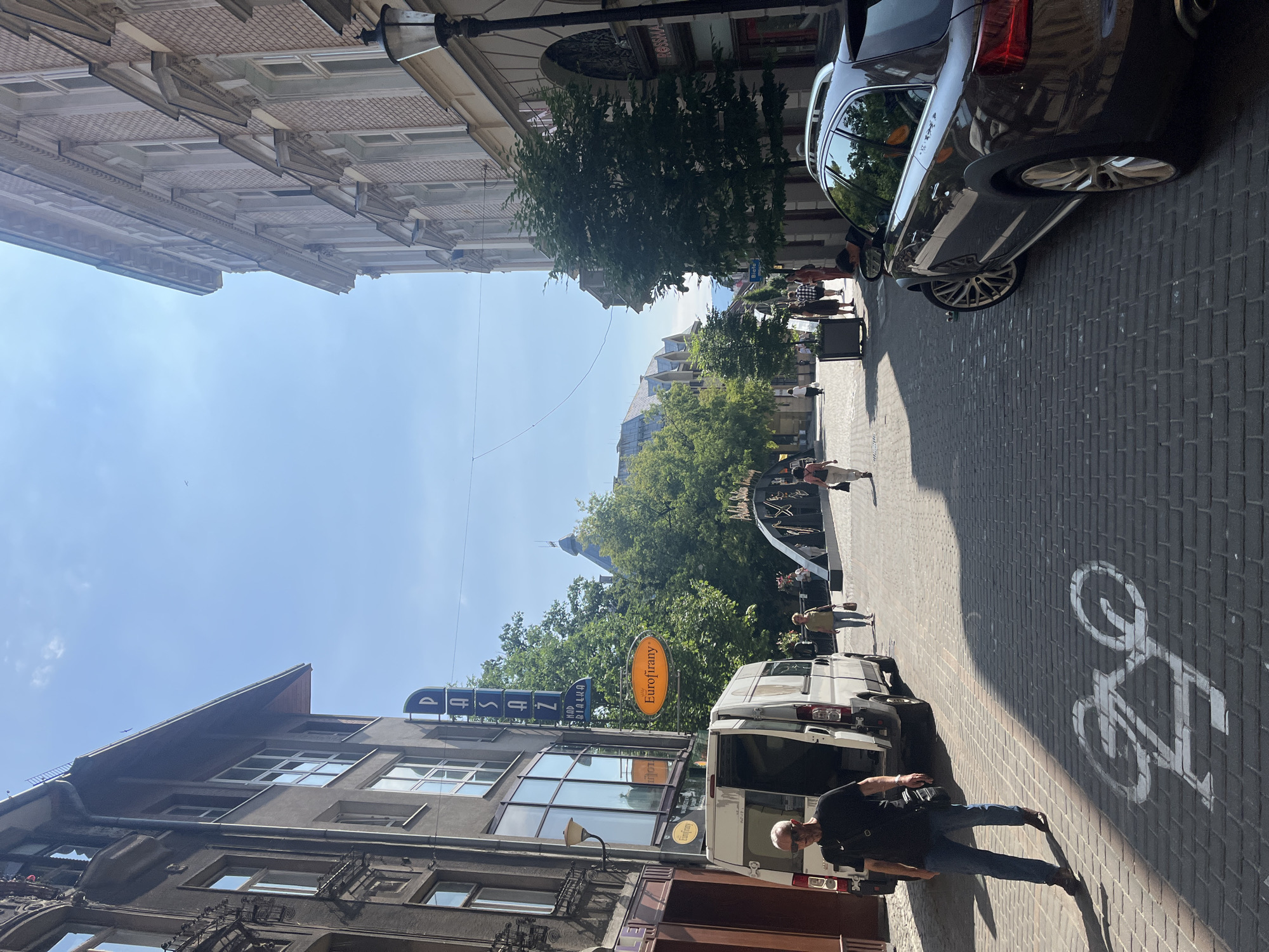 In a European city setting with cobblestone streets, a dark grey Audi station wagon is parked on the right side of the road, facing away from the camera. To its left, a white van is partially parked, with its rear door open and a man in a black t-shirt and blue jeans walking towards the viewer. The street is lined with buildings on both sides, with trees and an arched bridge visible in the background. A bicycle lane is marked on the street in front of the van. Several people are walking in the distance, and a street lamp is visible on the right. The sky is clear and blue.