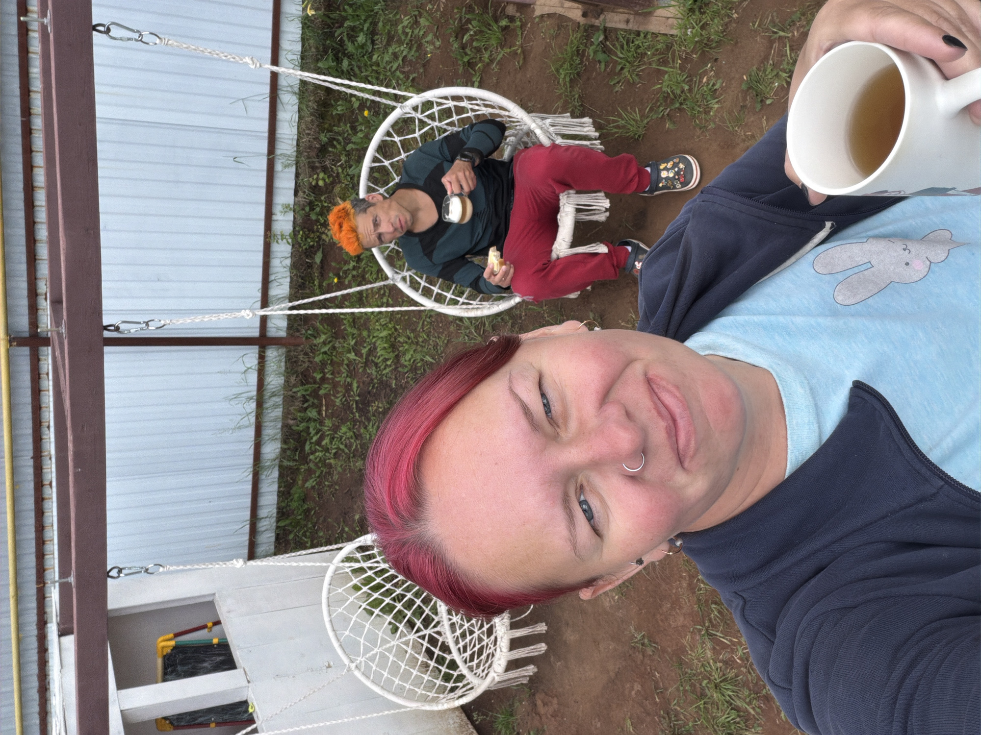In the city of Chaikovsky, Russia, two individuals are depicted outdoors in a backyard setting. In the foreground, a woman with bright pink hair takes a selfie, holding a white mug of tea. She is wearing a light blue t-shirt with a cartoon rabbit graphic and a dark blue zip-up jacket. Her left eye is partially closed as she smiles towards the camera, and she has a nose ring. Behind her, a man with spiky, bright orange hair is seated in a white macrame hanging chair. He is holding a piece of bread in his left hand and a glass of amber liquid in his right. He wears a dark long-sleeved shirt with black and dark green accents, red pants, and dark clogs with colorful patterns. Another empty white macrame hanging chair is visible to the left of the man. The background consists of a white corrugated metal fence, some greenery, and brown earth. A metal structure with yellow and brown beams is overhead, supporting the hanging chairs.