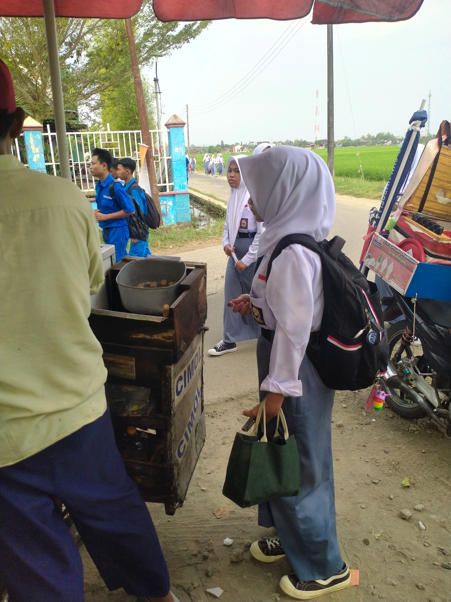 A street vendor is selling food from a cart in the city of Begajah, Indonesia. The cart is wooden and rusty, with a metal basin filled with food. A person in a light yellow shirt and blue pants stands in front of the cart, obscuring most of it. Behind them, two male students in blue uniforms are walking away from the cart. Further in the background, a group of female students in white hijabs and uniforms are walking on the road. The street is dirt and gravel, with some grass and vegetation on the side. A motorcycle is parked next to the cart, and a rice field is visible in the distance. The sky is overcast.