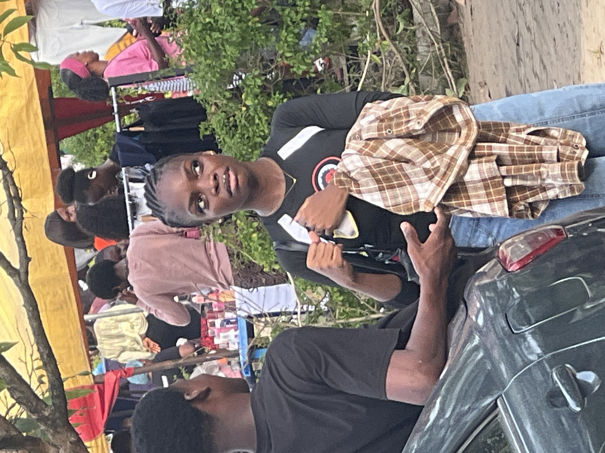 The image, taken in Ijanikin, Nigeria, captures a street scene with several individuals present. A young person wearing a black t-shirt with a logo, blue jeans, and holding a brown plaid jacket is facing the camera. They appear to be standing next to a dark-colored car, with another person in a black t-shirt partially visible beside the car. In the background, there are several other individuals near what seems to be a makeshift market or street vendor area. The environment includes trees, a yellow canopy, and various items of clothing and other objects displayed for sale.