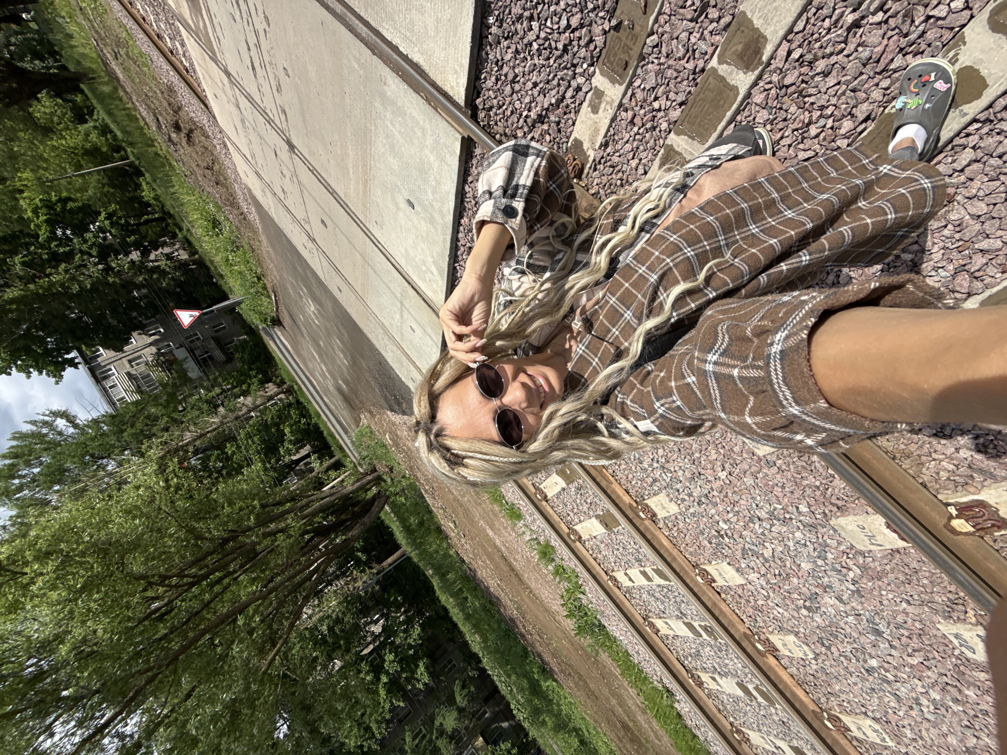 The image captures a selfie-style view of a woman lying adjacent to train tracks. She is wearing a brown plaid jacket and sunglasses. Her blonde hair is styled in braids. She appears to be positioned on the gravel and concrete adjacent to the tracks. On the left side of the image, there are trees, grass, and a building. The precise location appears to be a railway siding.
