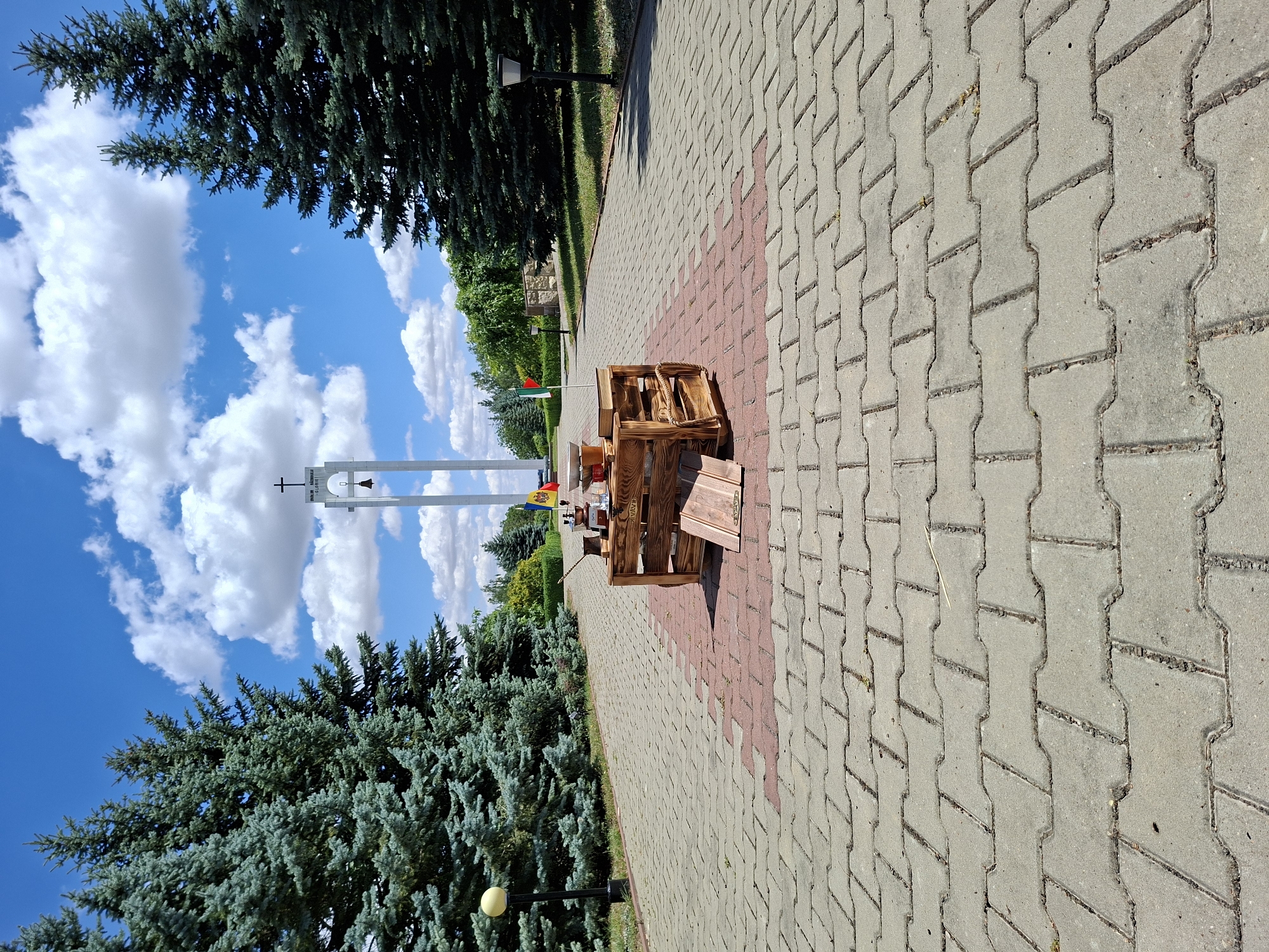 The image depicts an outdoor scene on a sunny day in Șerpeni, Moldova. In the foreground, a paved walkway made of interlocking concrete blocks leads towards a bell tower. The walkway has a pattern of red and grey blocks in the center. In the middle of the walkway, a wooden crate is set up, serving as a display. On top of the crate, there are several objects, including what appears to be a coffee making setup with a grinder and cups, two small flags, and a stack of books. One flag is red, yellow, and green, and the other is red and white with a green stripe. Further down the walkway, a tall, slender white bell tower with a cross at its apex is visible against a bright blue sky with scattered white clouds. The tower has an opening with a bell inside. Flanking the walkway are large, mature evergreen trees. In the background, there is more greenery and some low walls. The overall impression is that of a public space, possibly a memorial or a park, with a temporary setup for an event or display. There are no people visible in the image.