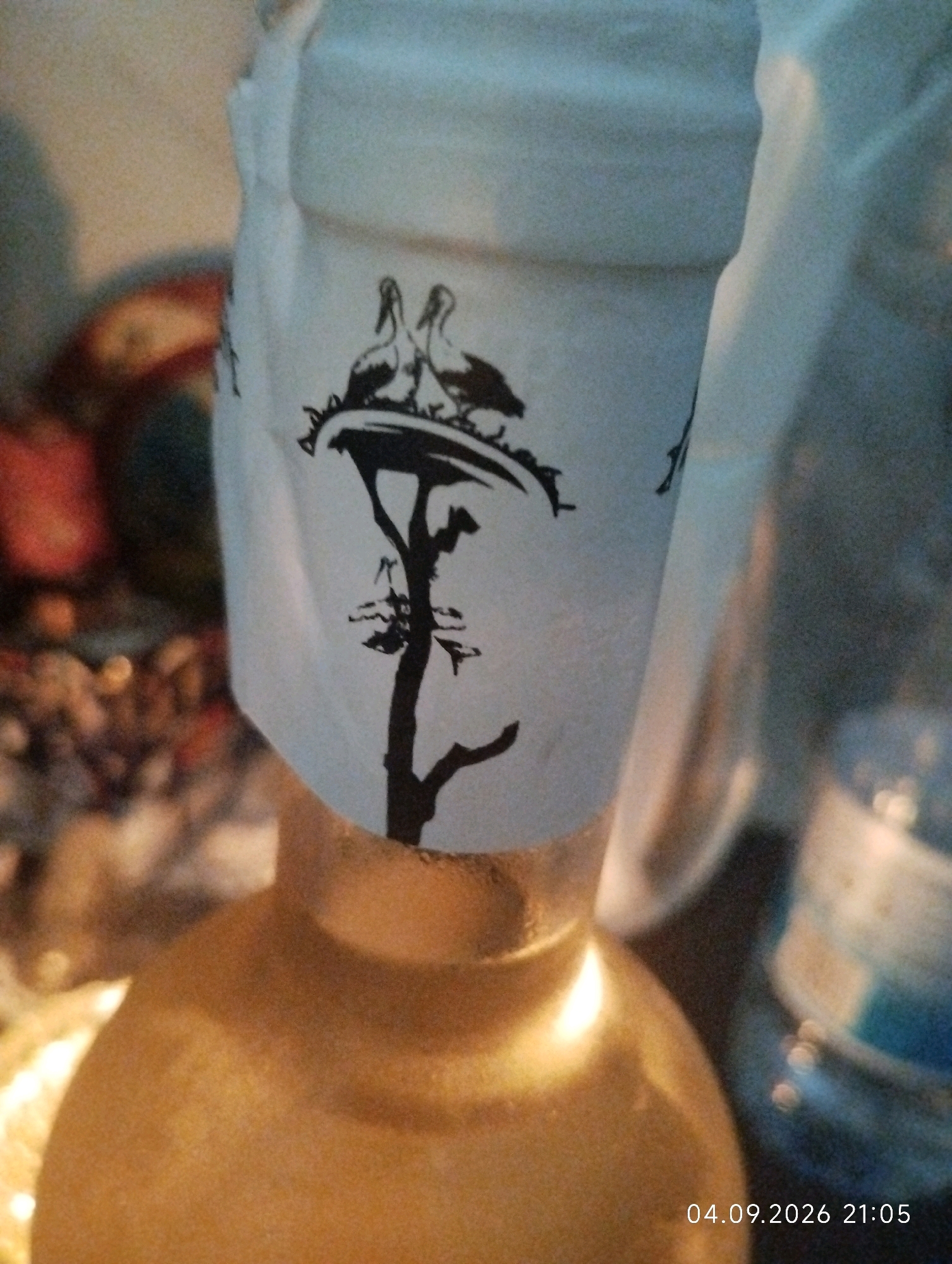 The image shows a close-up of the top of a bottle with a paper label. The label is white and features a black silhouette design of a tree with a nest containing two birds. The bottle appears to be golden or amber colored at the bottom. In the background, out of focus, are other objects, including what appears to be a red fruit, and a blue plastic bottle with some contents. The lighting suggests an indoor setting, possibly a home. A timestamp "04.09.2026 21:05" is present in the lower right corner. There is no visual information in the image that would indicate the location is the city of Ashkelon, Israel.