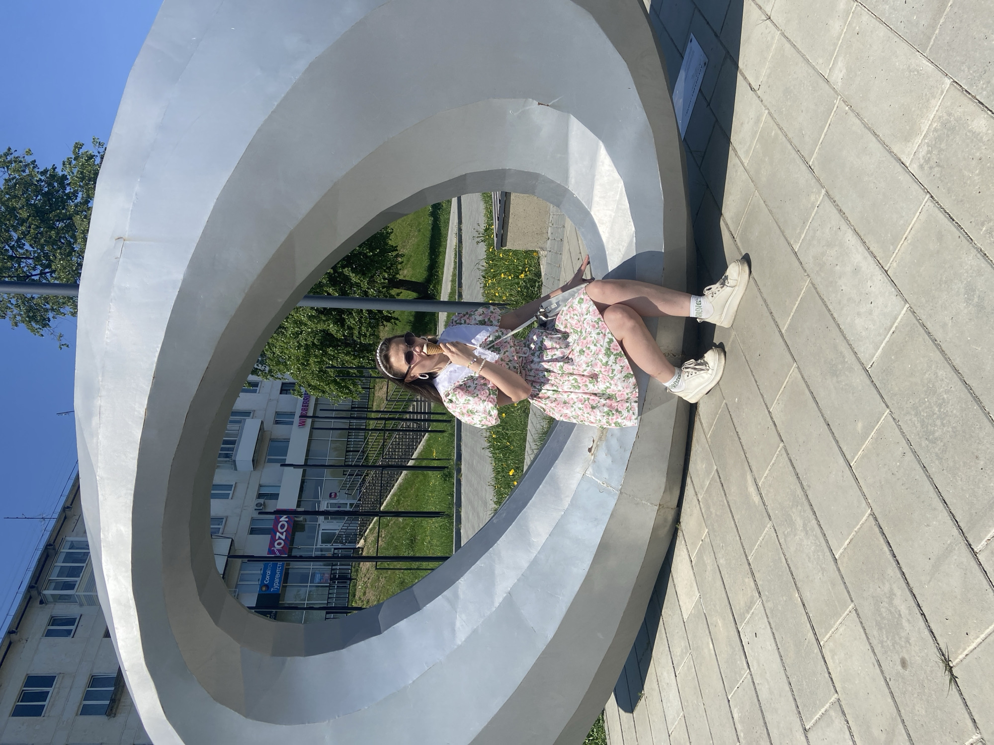 A young woman sits within a large, circular, silver-colored sculpture, eating an ice cream cone, in the city of Chaikovsky, Russia. The sculpture, with three concentric rings, is situated on a gray brick-paved area with a building visible in the background featuring an OZON logo. The woman is wearing a floral dress, white sneakers, sunglasses, and a white headband. She holds the ice cream cone in her right hand, near her face. Green grass, trees, and a black metal fence are visible through the sculpture. The sky is clear and blue.