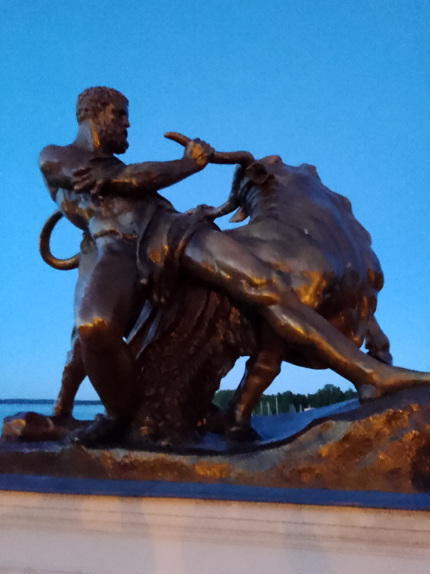 This is a low-angle, medium shot of a bronze sculpture depicting a muscular man wrestling a bull. The sculpture is illuminated by artificial light, highlighting the metallic sheen and details of the figures. The man, shown from the waist up, is on the left, gripping the bull's horns with his right hand and its muzzle with his left. His body is contorted in a powerful struggle, with his legs braced against the bull's body. The bull, facing to the right, is also depicted in a dynamic pose, its body angled and its head turned, appearing to resist the man's efforts. The sculpture is mounted on a textured, rocky base, which sits on a darker, possibly stone or concrete, platform.
In the background, the sky is a deep, clear blue, suggesting either twilight or early morning. A faint horizon line indicates a body of water, likely a lake or bay, with a distant tree line and some structures, possibly boats or buildings, visible along the shore. The overall mood is one of intense struggle and raw power. The style of the sculpture is realistic, with attention to musculature and anatomical detail.
Given the location context of Schwerin, Germany, this sculpture may be related to mythology or a historical event. The scene evokes the myth of Hercules and the Cretan Bull, or perhaps a depiction of the Taurus constellation. The light sources, likely artificial streetlights or floodlights, cast a warm glow on the bronze, creating strong highlights and shadows, enhancing the dramatic effect of the composition. There is no visible text in the image.