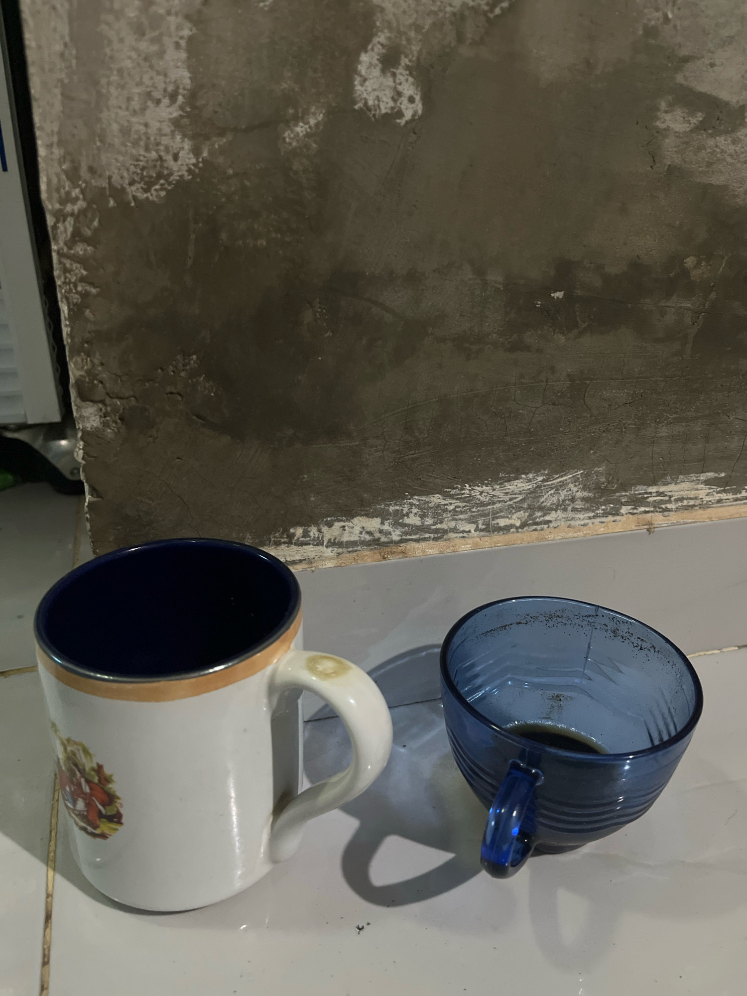 Two mugs are on a tiled floor, against a rough, grey, unfinished concrete wall. One mug is white with a blue interior and a decorative image on its side. The other mug is blue glass, with a small amount of dark liquid at the bottom. A shadow from the blue mug extends to the right. The scene is indoors, likely in an unfinished area of a building, possibly a residential or commercial space in Kebumen, Indonesia.