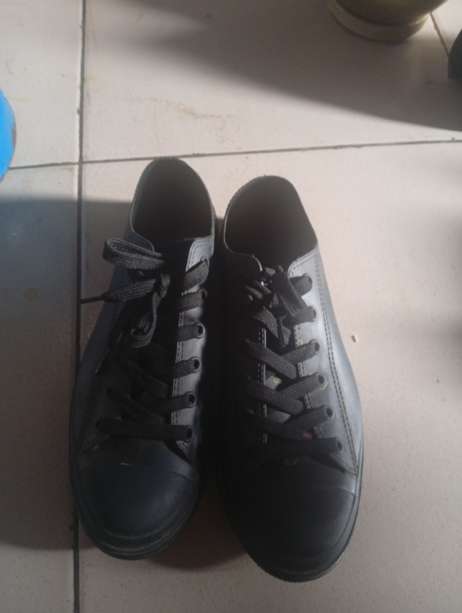 A pair of black, low-top lace-up sneakers is shown resting on a tiled floor. The shoes are positioned side-by-side, with the laces tied in bows. The floor is composed of light-colored square tiles with visible grout lines. The lighting suggests an indoor environment, possibly a room or hallway. The specific location in Ashata, Nigeria, is not discernible from the image itself.
