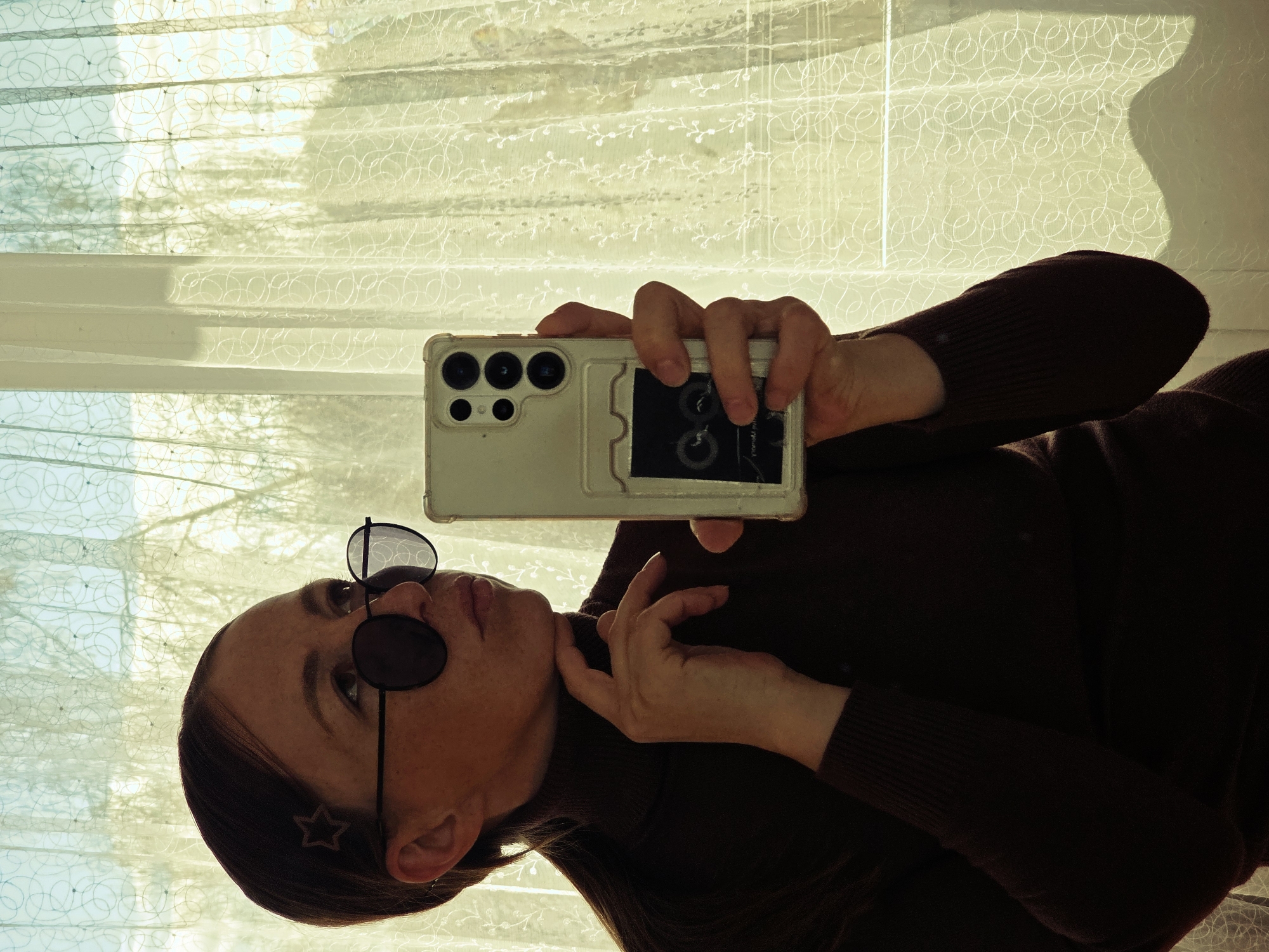 A person wearing dark sunglasses and a brown turtleneck is holding a smartphone in front of a window covered with translucent curtains. The person's face is turned towards the camera, and they are touching their face with one hand. The smartphone case is light-colored and has a small, dark object attached to the back. The window is letting in bright sunlight, and the curtains have a decorative pattern. The location is likely an indoor space in the city of Chaikovsky, Russia.
