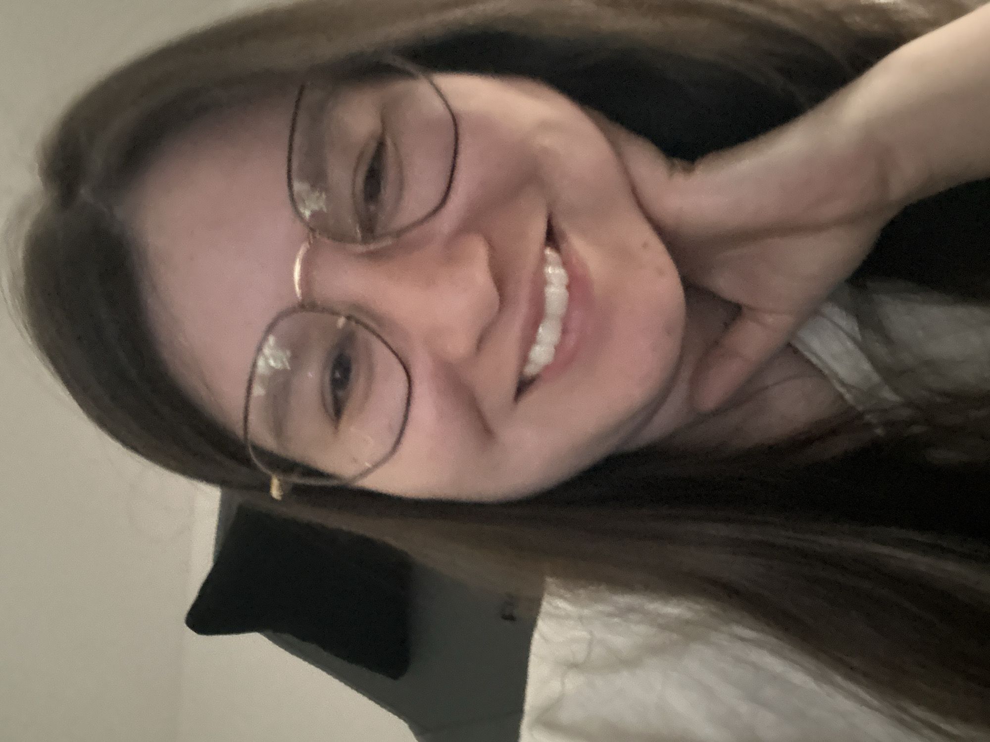 A woman with long brown hair and glasses is smiling. She has fair skin and is wearing a light-colored shirt. Her hand is near her face. The background is blurred, showing a headboard and wall. The image was not captured in Câmara de Lobos, Portugal. There is no evidence of the city, region, or country within the image.
