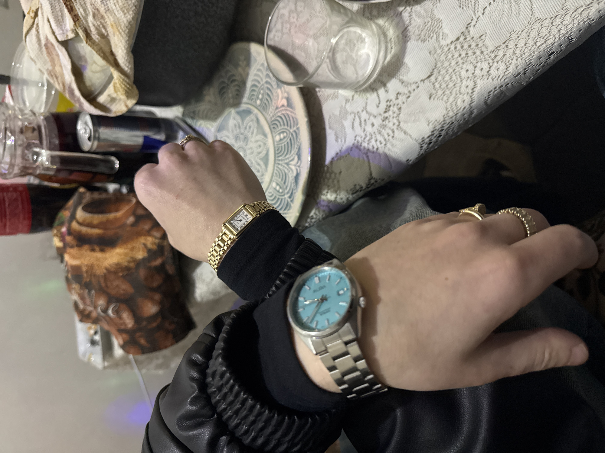 The image shows two hands wearing watches. The left hand has a gold watch with a white face and a gold band. The right hand has a silver watch with a light blue face and a silver band. Both hands also have rings on the fingers. The hands are positioned on a table covered with a lace tablecloth. In the background, there are several objects, including a plate, a glass, a can, and a decorative object with an animal print. The environment suggests an indoor setting. There is no information in the image to indicate it was taken in Yerevan, Armenia.