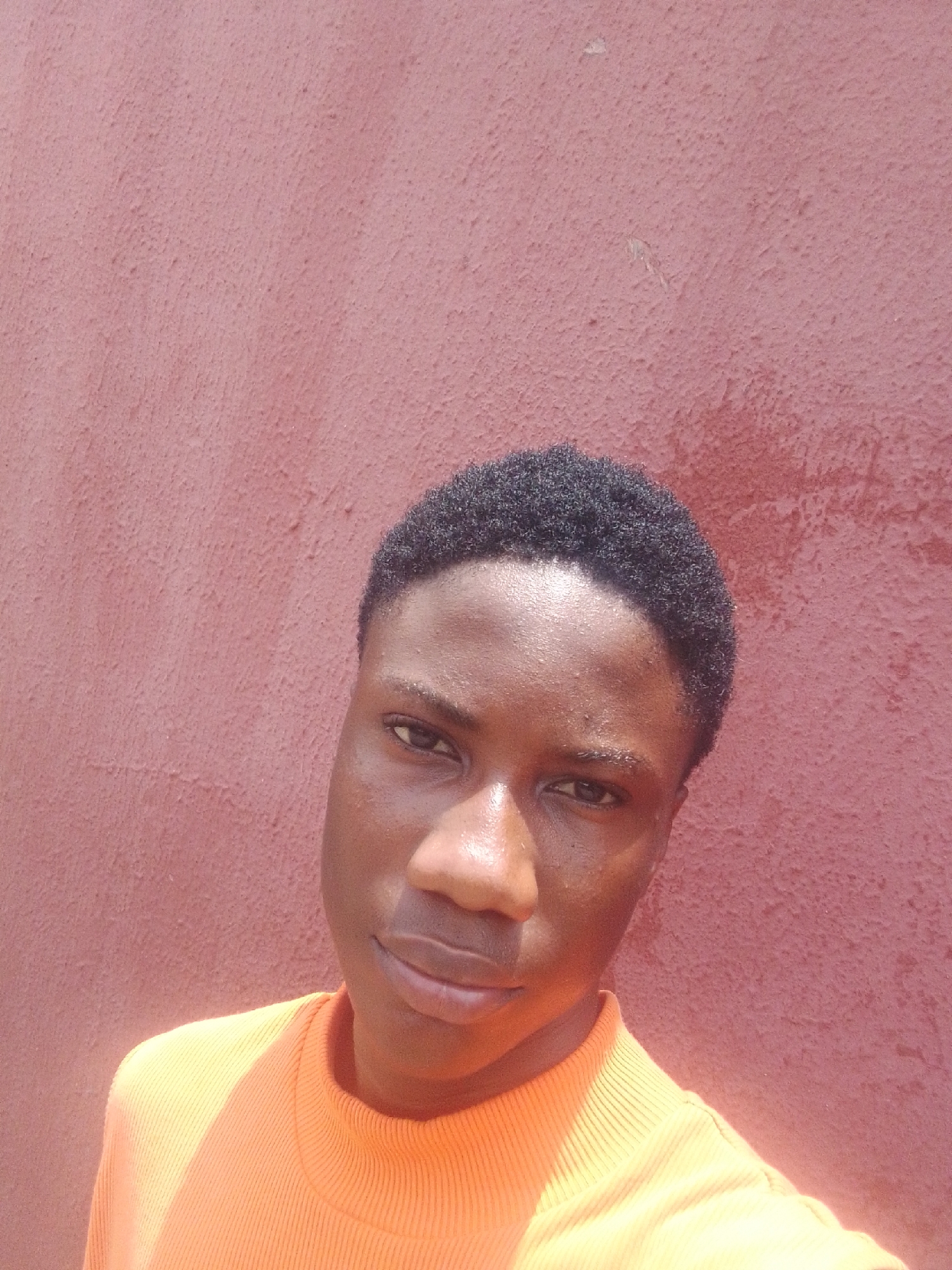A young, dark-skinned person with short, dark hair, wearing an orange ribbed sweater, stands in front of a textured, reddish-brown wall. The individual is looking directly at the camera with a neutral expression. The lighting suggests an outdoor setting, potentially in Onitsha, Nigeria, due to the likely location of the requester. The background appears to be a simple, unadorned wall, common in urban environments. No other subjects or objects are visible in the immediate foreground or background. The person is alone.