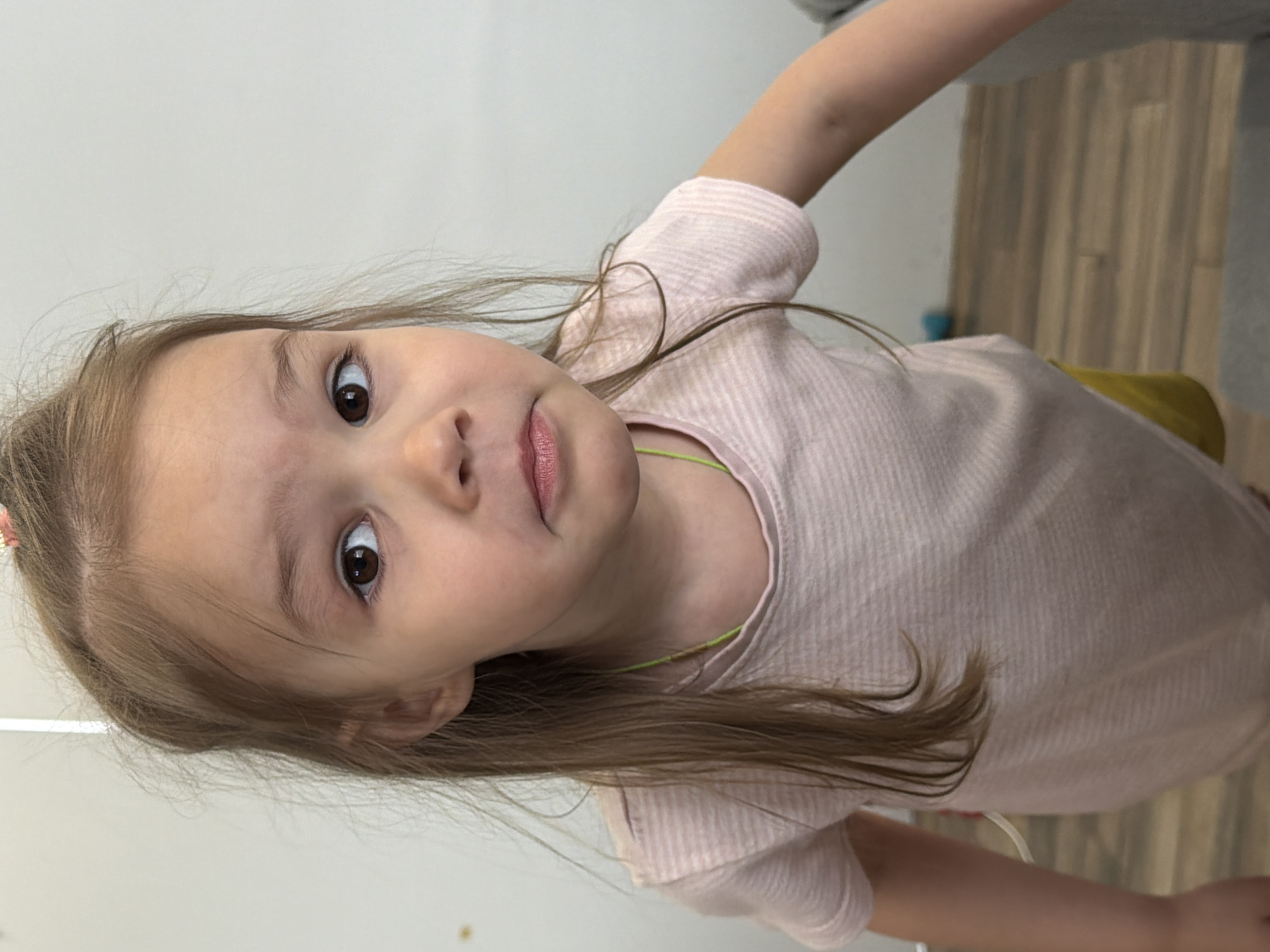 A young girl with light brown hair and brown eyes is the focus of the image. She is wearing a light pink, striped t-shirt and a lime-green necklace. The background includes a white wall, wood-patterned flooring, and a grey sofa. The location cannot be identified as Chaikovsky, Russia, based on the visual information provided.