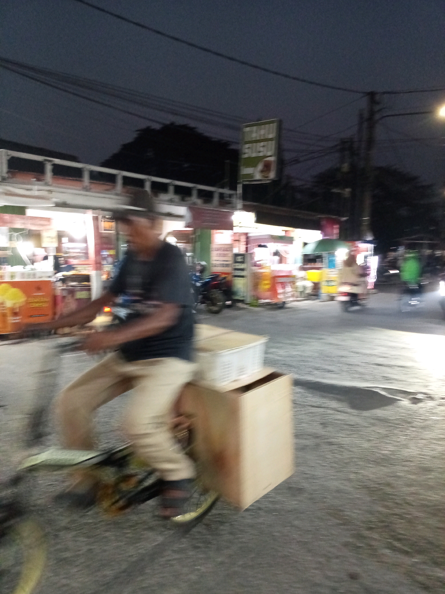A blurry, motion-streaked image captures a street scene at dusk or night in Bekasi, Indonesia. In the foreground, a person is riding a bicycle with a large cardboard box attached to the rear. The rider, wearing a dark t-shirt and light-colored pants, is in motion, as indicated by the blur. The bicycle has a visible wheel with spokes. Behind the bicycle, a white plastic container is also visible.
The background features a row of food stalls or shops, some illuminated with bright lights, suggesting a commercial area. A sign with green lettering, possibly advertising "Tahu Susu," is partially visible above the stalls. Several motorcycles are parked or in motion, and streetlights illuminate the asphalt road. The overall impression is that of a busy street at night with vendors and activity. The motion blur suggests the photograph was taken from a moving vehicle or with a slow shutter speed.