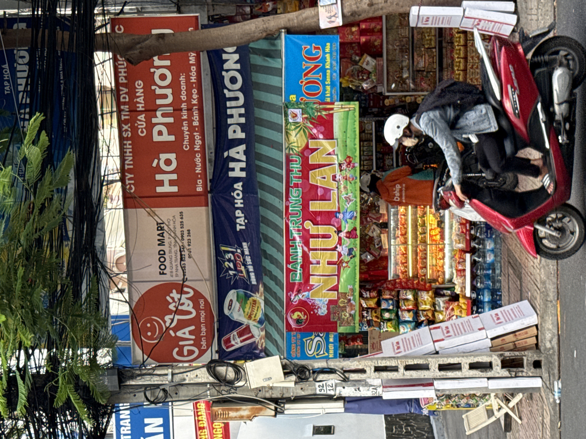The image depicts a street scene in Nha Trang, Vietnam, with a focus on storefronts and a street vendor. A red scooter with a rider wearing a white helmet and denim jacket is parked in the foreground, partially obscuring a storefront. The storefronts display various signs, including "FOOD MART Gia Tot" and "Cửa Hàng Hà Phượng," indicating a grocery store and a general store. The shelves inside the stores are stocked with an assortment of packaged goods, including snacks, beverages, and other consumer products. A person in an orange jacket is visible inside one of the stores, potentially working as a vendor. The environment appears to be a bustling urban area, with electrical wires strung overhead and other businesses visible in the background. The overall impression is one of everyday commerce in a Vietnamese city.