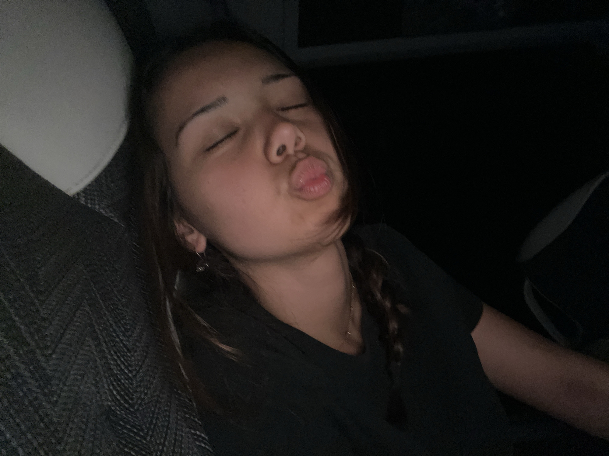 A young woman is resting in what appears to be a car seat. Her eyes are closed, and her lips are pursed in a kissing gesture. She has dark hair styled in braids and is wearing a dark shirt. The surroundings are dark, with the headrest of the car seat visible. The location is Unknown, Unknown.