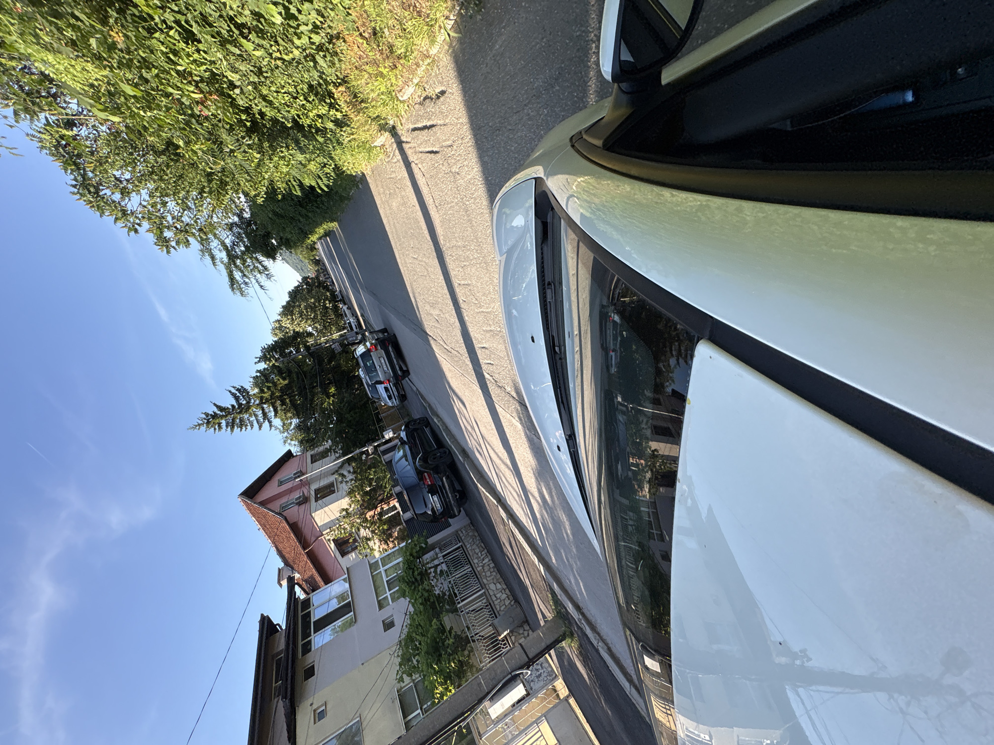 The image captures a street scene in Vratsa, Bulgaria, on a sunny day. The perspective is from inside a white car, looking forward and to the left. On the left side of the street, there are several residential buildings, some with red-tiled roofs, and a white fence. A black SUV is parked on the left side of the road. On the right side of the street, there is a dense green hedge lining the road. The road itself is paved and has distinct shadows cast by trees and possibly buildings. The sky is clear blue with a faint contrail visible. Several other cars are seen parked and driving further down the street. The overall environment suggests a residential neighborhood with some vegetation.