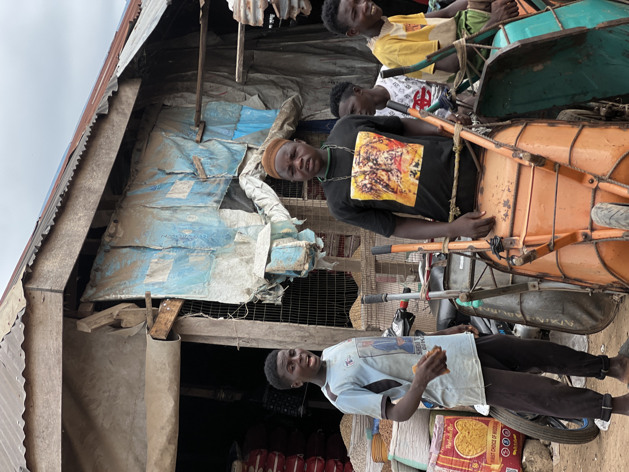 The image depicts a scene in Oko Amakom, Nigeria. Three young men are visible, two of whom are positioned behind a cart carrying two large drums, one orange and one teal. The man in the foreground on the left, wearing a black t-shirt with a colorful graphic and a brown cap, looks towards the camera with a somewhat strained expression. He has a chain around his neck. To his right, another young man, wearing a light blue t-shirt with a graphic and dark pants, is holding a piece of food in his right hand and looking to the side. His mouth is open, suggesting he might be speaking or in the process of eating. Behind these two, a third young man, wearing a yellow t-shirt, is visible, looking away.
The setting appears to be a market or roadside stall, with makeshift structures made of corrugated metal and tarps. Bags of what appear to be grains or rice are stacked on the ground. The overall atmosphere suggests a daily life scene, possibly during daylight hours given the illumination. There are no clear indications of weather, but the sky is overcast. The cart with the drums and the surrounding market goods suggest an active commercial environment. The expressions on the faces of the young men are varied, with the one in the black shirt looking directly at the camera, the one in the blue shirt engaged in eating and speaking, and the one in the yellow shirt looking away. The image captures a slice of everyday life in this Nigerian locale. Text is visible on the orange drum, partially obscured, and on a bag of rice in the lower right corner, which reads "Commodity Rice" and "PULOTAR RICE".