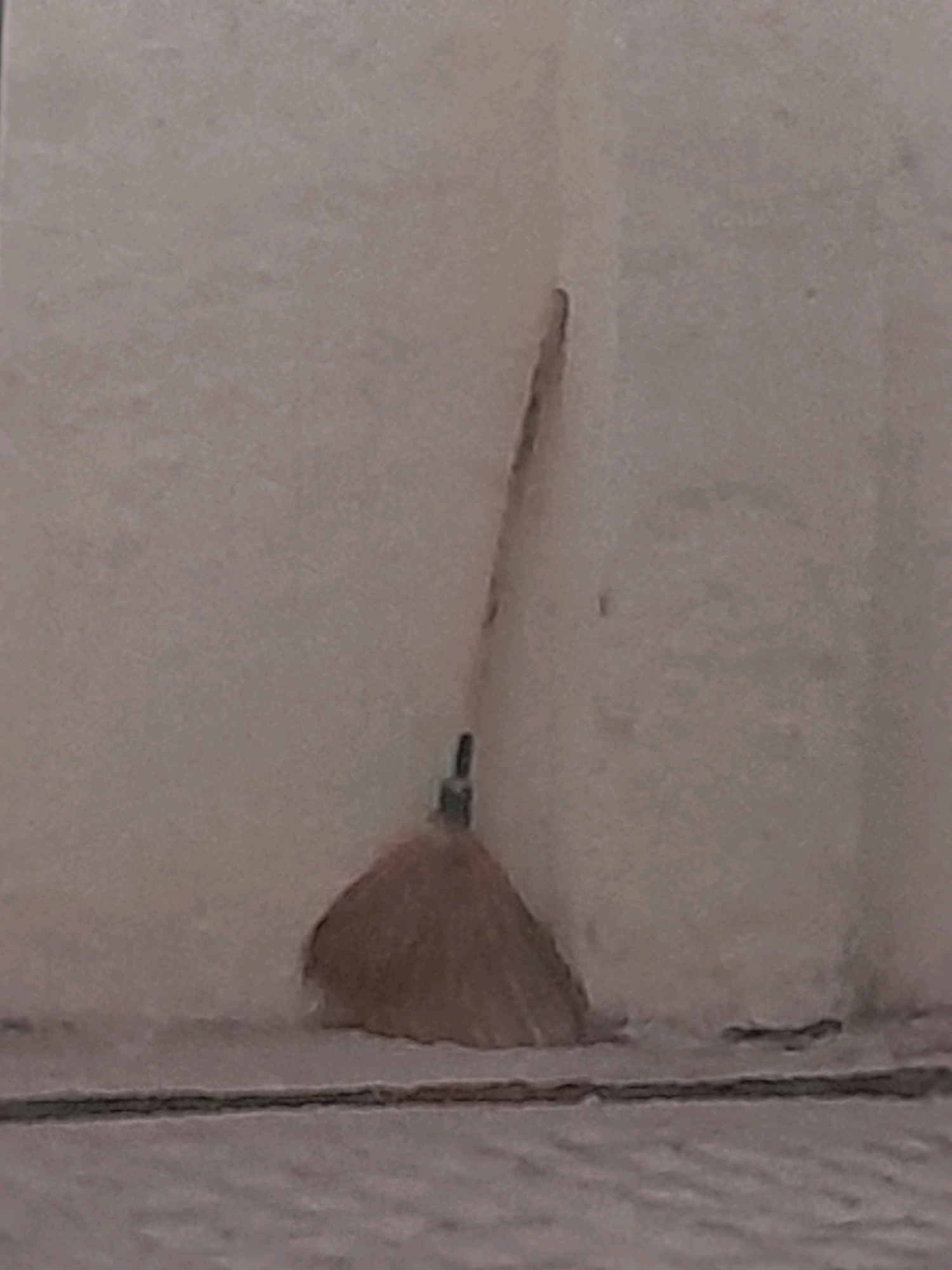 An out-of-focus, slightly blurry photograph shows a light brown broom leaning against a pale beige wall in the corner of a room. The broom has a dark handle and a fanned-out brush made of what appears to be straw or dried plant material. The wall is textured and shows some minor discoloration and subtle marks. A thin dark line, possibly a baseboard or the edge of flooring, runs horizontally across the bottom of the image. The floor itself is a lighter, mottled gray color. The overall lighting is dim and diffused.
