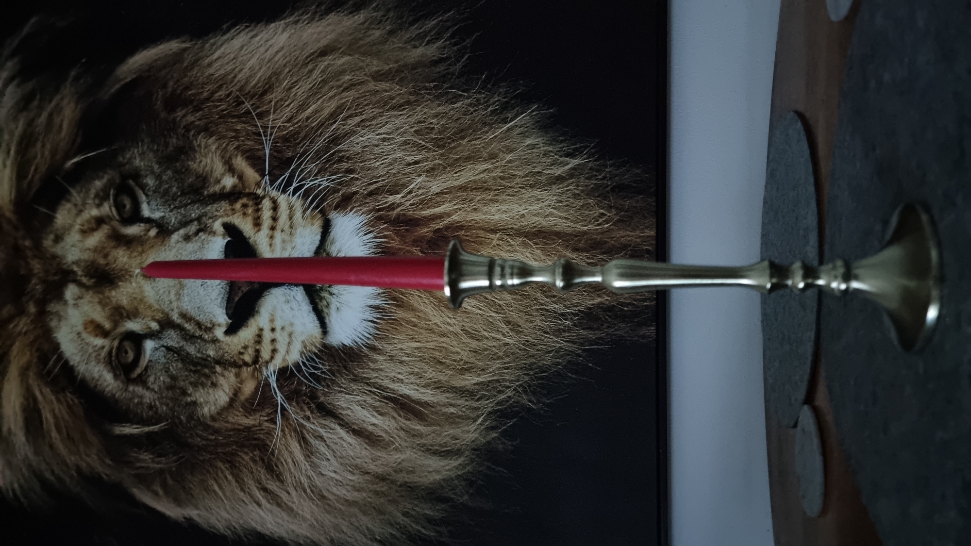 A red candle stands upright in a brass candlestick, positioned in the foreground of the image. The candle is centered and appears to be the primary subject. Behind the candlestick and candle is a framed artwork depicting the face of a lion. The lion's mane is prominent and detailed, taking up a significant portion of the background. The artwork is mounted on a wall, the top edge of which is visible in the upper portion of the image. The candlestick rests on a table surface, partially obscured by what appear to be two circular placemats or coasters. The lighting is dim, casting shadows and creating a somber atmosphere. The scene suggests an indoor setting, likely a home environment. There is no indication of the city of Heidenheim an der Brenz, Germany.