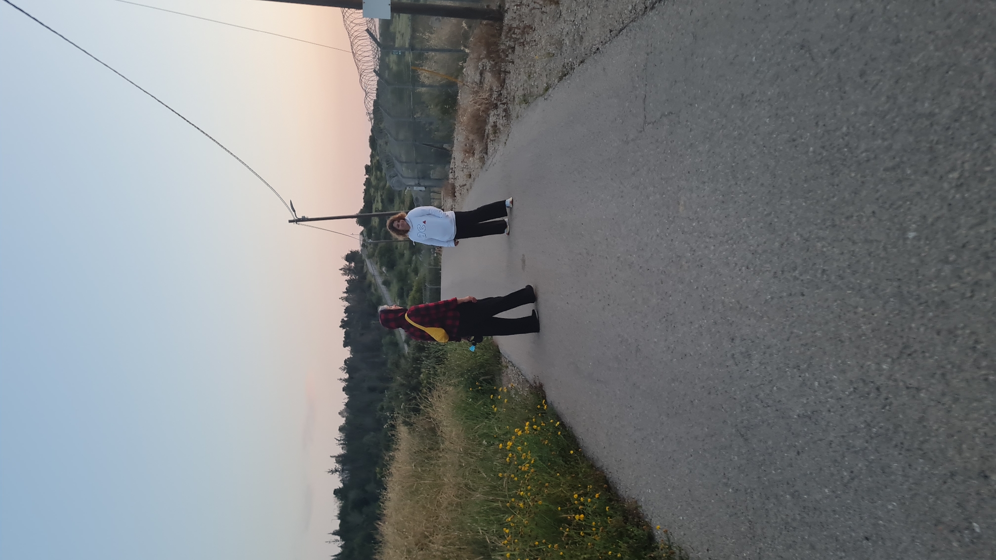 Two people stand on a paved road in the Lachish Regional Council, Israel. One person wears a red and black plaid shirt and a yellow bag. The other person wears a white sweatshirt. A fence with barbed wire runs along the side of the road. Vegetation including yellow flowers grows on the side of the road. Power lines stretch overhead. The sky is light blue and pink, suggesting dawn or dusk.