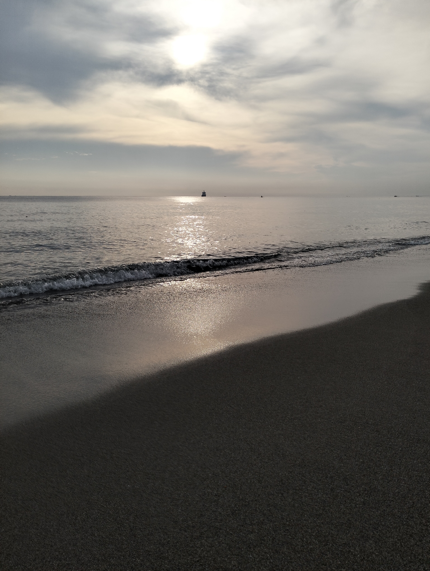 The image shows a beach scene in Lhokseumawe, Indonesia. The sky is overcast with the sun obscured by clouds. The sea is calm with small waves lapping the shore. The beach has a dark sand, reflecting the light from the sky. There is a ship in the distance on the horizon.