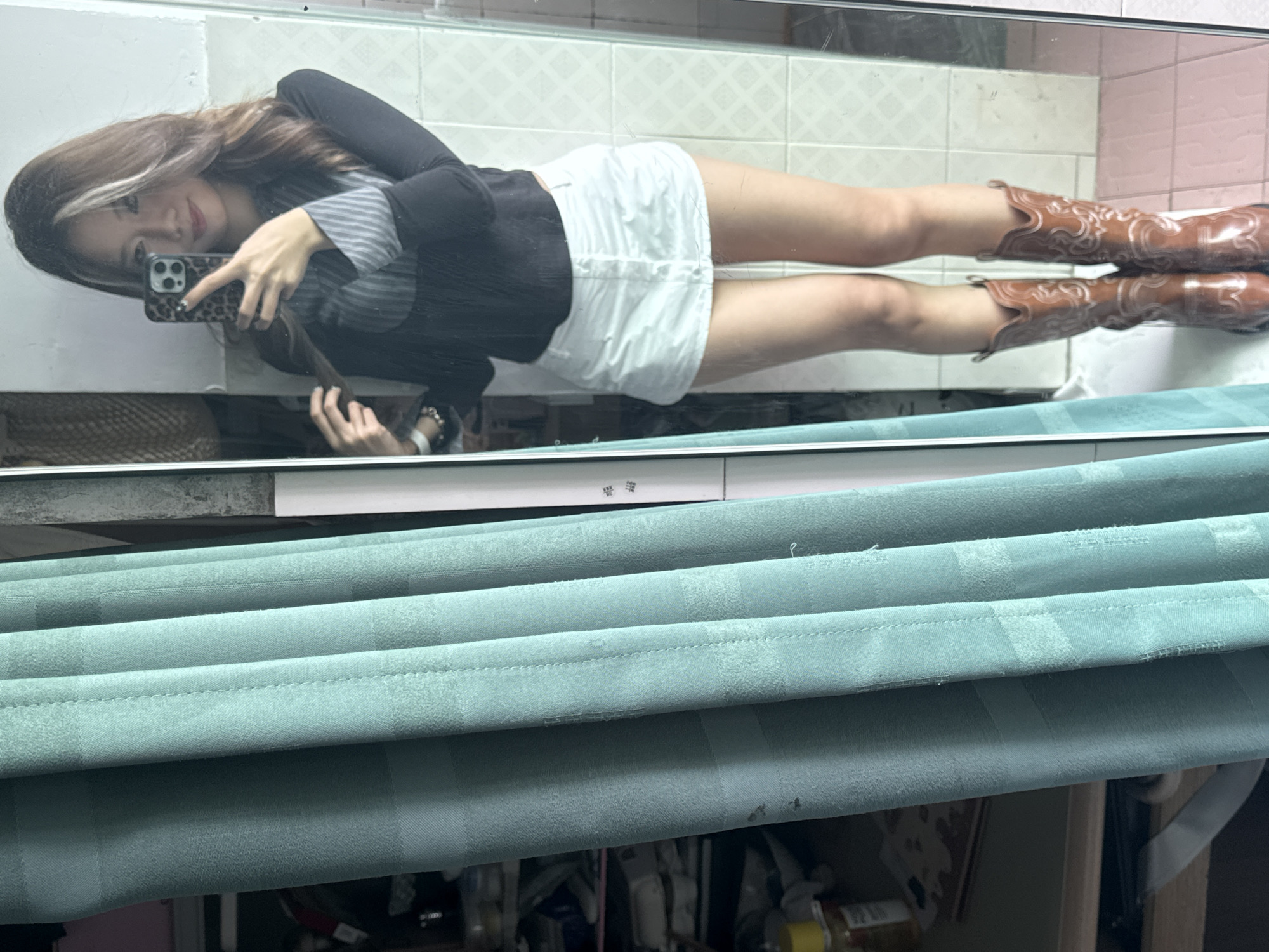 A young woman takes a selfie in a bathroom mirror. She wears a striped shirt under a black cardigan, a short white skirt, and brown cowboy boots. The bathroom has tiled walls. In the foreground are light blue curtains. The location is likely in Nan'an District, China, but this is not directly visible in the image.