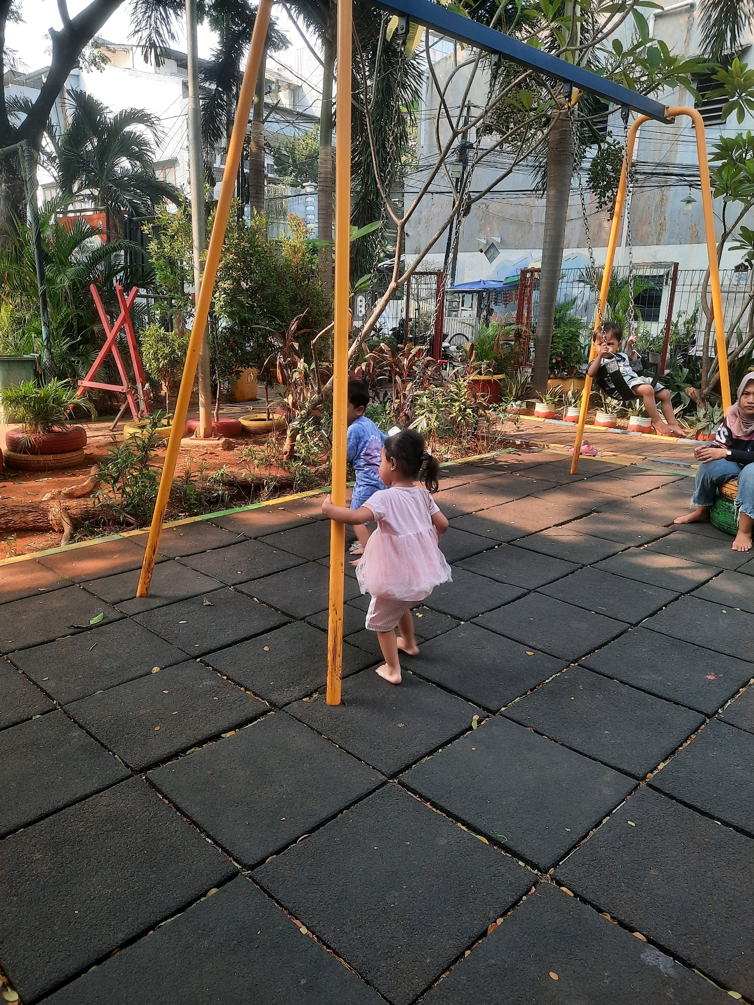 A group of people are at a playground with a swing set. A girl in a pink dress is walking on the rubber matting. A boy in a blue shirt is on the swing. A woman in a hijab and jeans is sitting on a tire. The playground is surrounded by lush green plants and trees, with buildings in the background. The location is the Taman Impian Jaya Ancol in North Jakarta, Indonesia, a popular tourist attraction known for its beach, theme park, and aquarium.