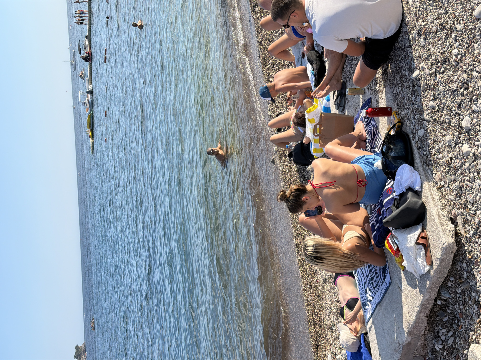A group of people are relaxing on a pebble beach in Budva, Montenegro. The sea is calm and blue, with several boats in the distance. On the beach, people are sunbathing on towels, some are in the water, and others are sitting and talking. There are bags, bottles, and other beach accessories scattered on the sand. The sun is shining, and the sky is clear.