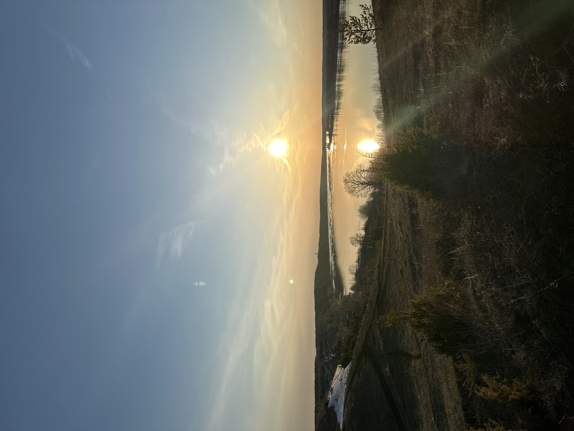 The image depicts a horizontal view of a landscape at sunset in Ольховка, Russia. The sky is a gradient from blue to orange with light clouds scattered throughout. The sun is a bright, orange disc reflected in the calm water of a river or lake. The landscape on the right is a mixture of vegetation, trees, and brown, dry grass. A village with some snow is visible on the left, at the bottom of a hill. The image is slightly tilted.