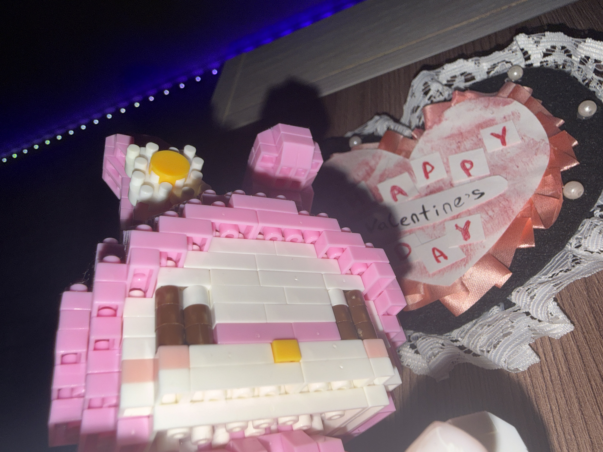 A pink and white block-built character, resembling a cat, is positioned on a wooden surface. The character has brown eyes and a yellow square on its muzzle. To the right of the character is a heart-shaped decoration with "HAPPY VALENTINE'S DAY" written on it in red letters on white squares. The decoration is bordered by pink ribbon and white lace, with small white pearls attached. In the background, a string of purple lights is visible against a dark surface. There are no people visible in the image. The scene is lit by an artificial light source, casting shadows. The location is not specified in the image or its metadata.