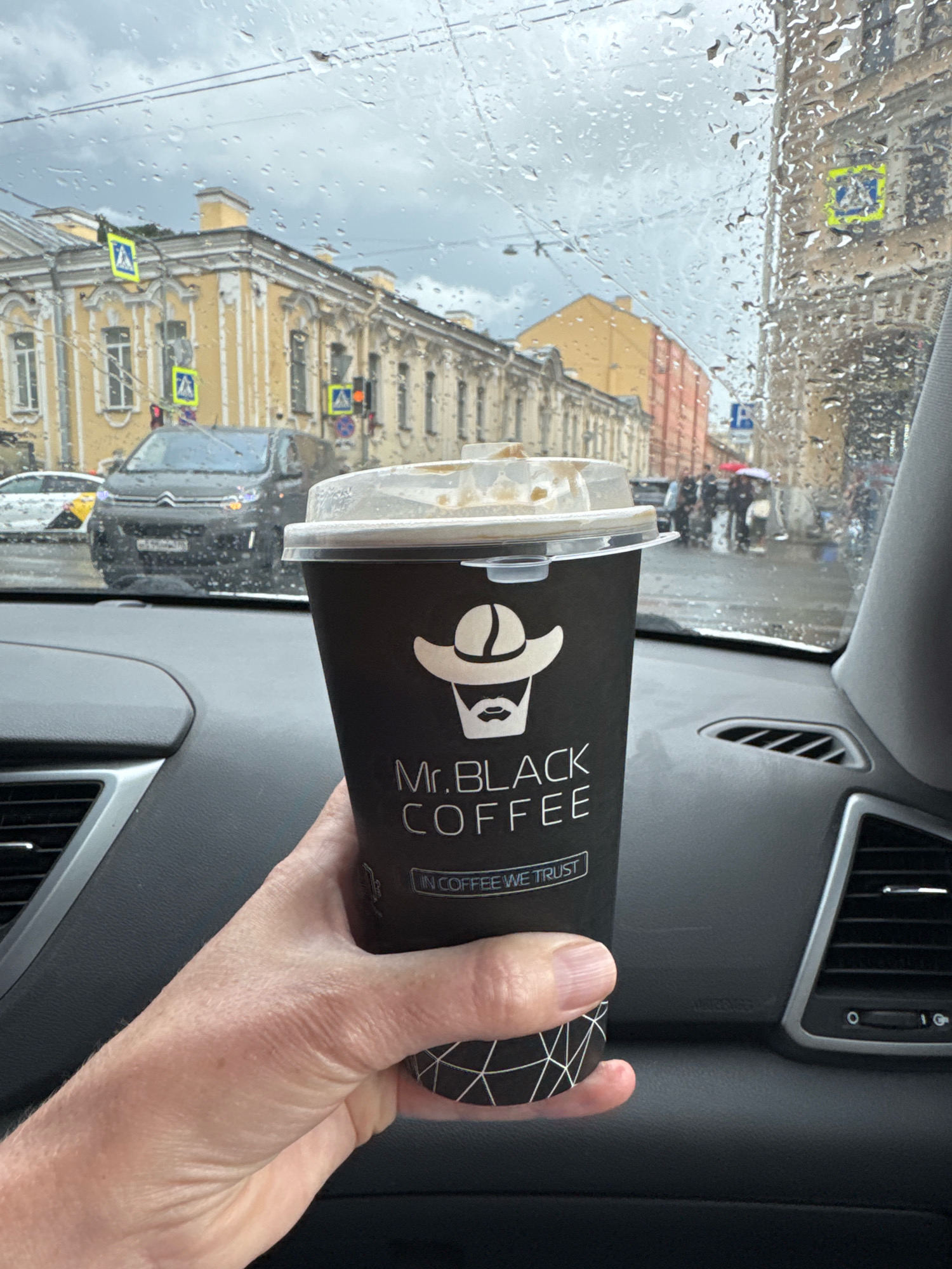 A hand holds a coffee cup inside a car, with the windshield showing a rainy city street in Saint Petersburg, Russia. The cup is black with the "Mr. BLACK COFFEE" logo, featuring a stylized cowboy hat and beard. Outside, buildings with ornate facades line the street, and pedestrian crossing signs are visible. Cars are present, and people with umbrellas are walking in the rain. The dashboard of the car is visible in the foreground.