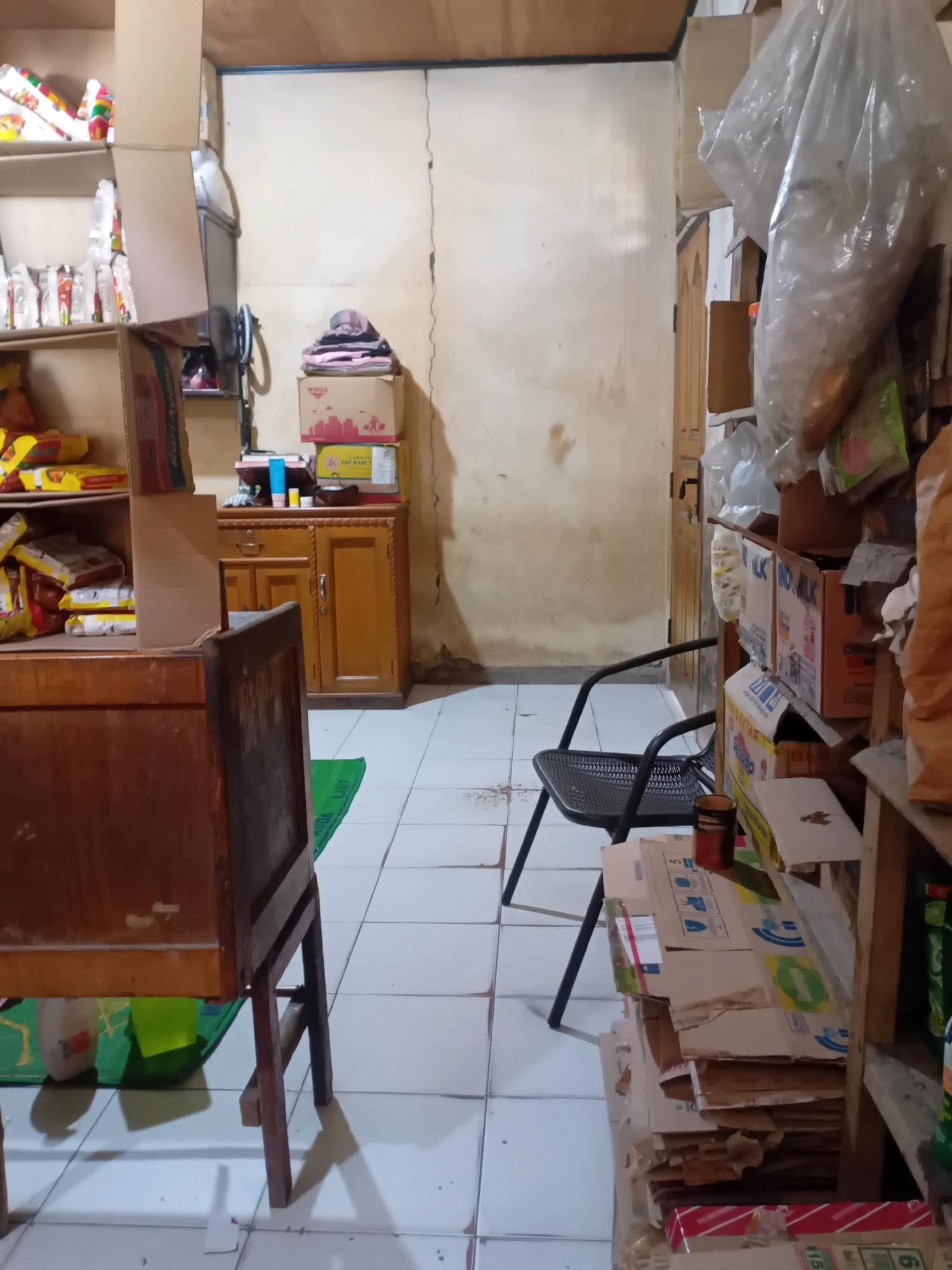 This is an indoor scene that appears to be a storage area or a small shop in Pekanbaru, Indonesia. The room has tiled flooring and plain, possibly plastered walls that show some discoloration and cracking. On the left side of the image, there are several shelves stacked with various items, including what appear to be food products in packaging. A portion of a wooden desk or table is visible in the foreground, partially obscuring the lower part of the shelves. To the right of the shelves, there is a wooden cabinet with drawers and doors. Further to the right, a black metal chair is positioned. Behind the chair, there are more shelves filled with boxes, plastic-wrapped items, and other assorted goods. A tall wooden door is visible in the background on the right side. The overall impression is one of clutter and organized storage of merchandise.