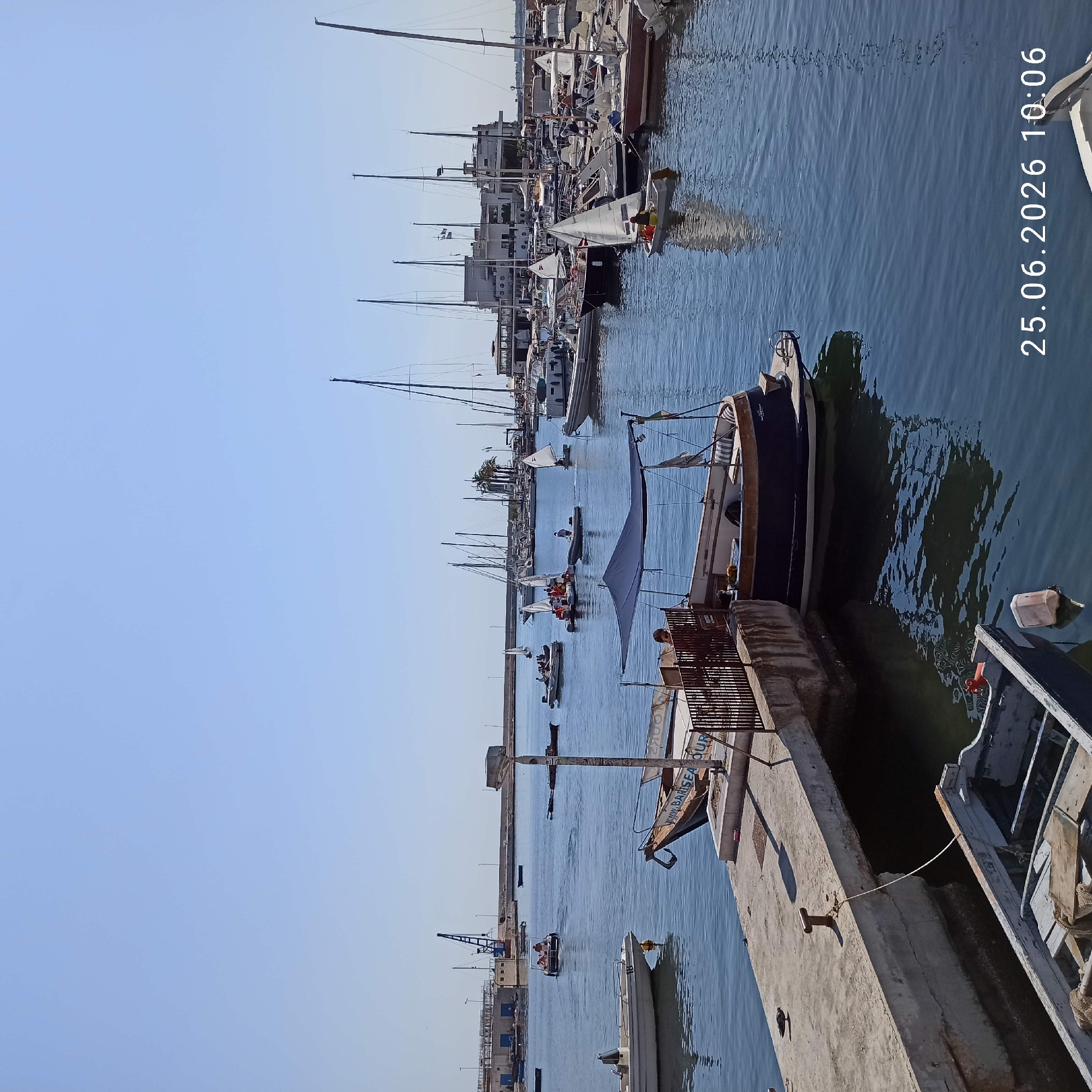 This is a vertical, eye-level medium shot of a marina with numerous sailboats and smaller boats docked and in the water. The sky is a clear, pale blue. The water is calm and reflects the light.
In the foreground, a concrete pier runs along the right side of the frame. A weathered, wooden boat is tied to the pier, and another boat, partially obscured, is also docked. Further down the pier, a metallic grate structure is visible, with a person standing behind it.
In the mid-ground, several small sailboats with white sails are in the water. Some appear to be occupied by people, possibly engaged in sailing activities. Larger sailboats are docked further into the marina. Buildings, some multi-story with balconies, line the shore behind the docked boats.
In the background, more sailboats are visible, along with what appears to be a crane and other industrial structures. Palm trees are also scattered amongst the buildings.
A date stamp, "25.06.2026 10:06," is visible in the upper right portion of the image, suggesting the time of day is around mid-morning. The overall impression is a sunny day at a lively marina.