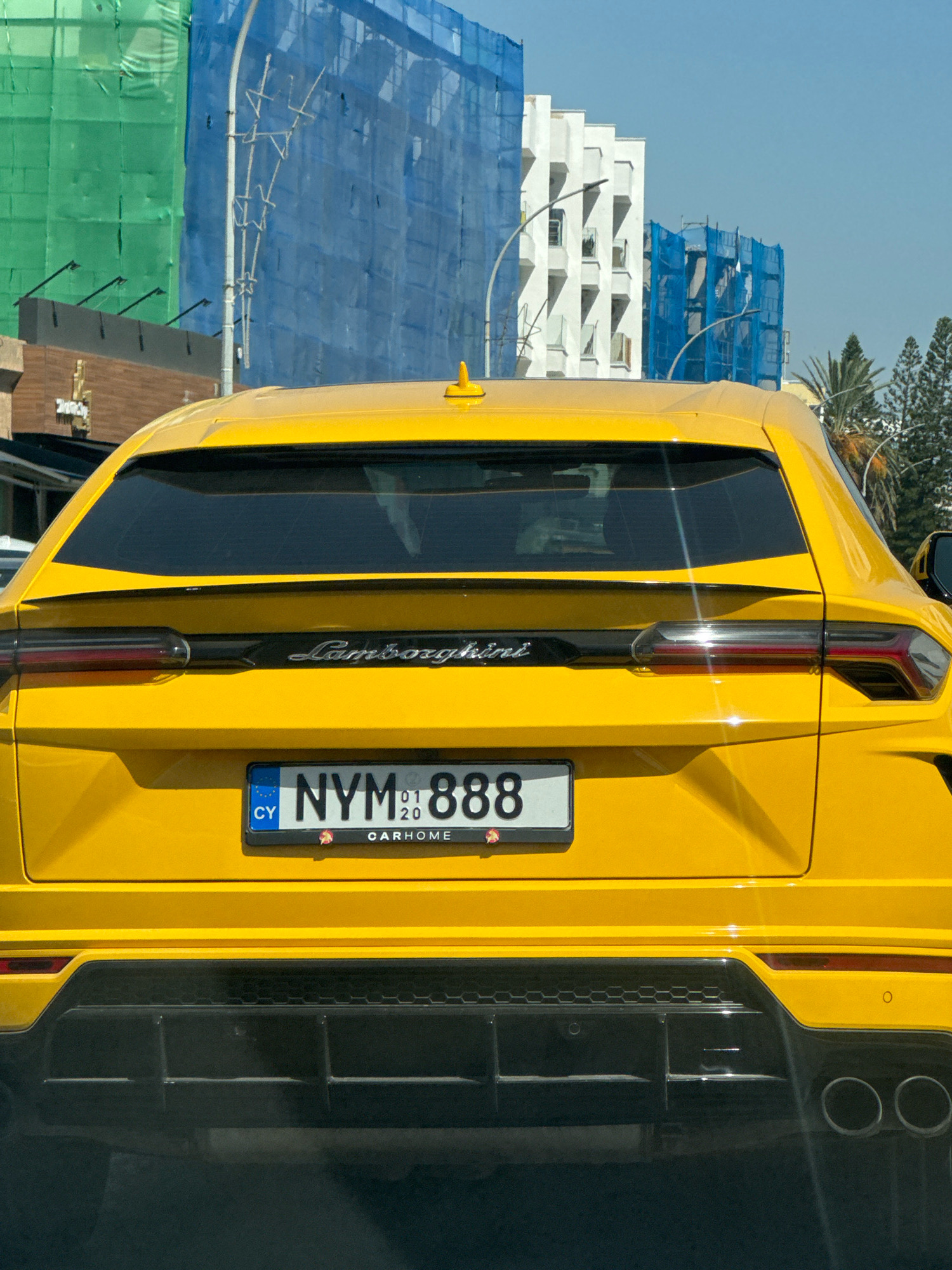 A yellow Lamborghini Urus is parked on a street in Germasogeia Municipality, Cyprus. The car is viewed from the rear, with its license plate prominently displayed. The license plate reads "NYM 888" and also shows "01 20" and "CARHOME". The car is a sports utility vehicle with a distinctive rear spoiler and quad exhaust pipes.
The background of the image shows a building under construction, covered in blue and green scaffolding nets. The sky is clear and blue. The scene appears to be a sunny day in an urban or semi-urban environment. No people are visible in the image.