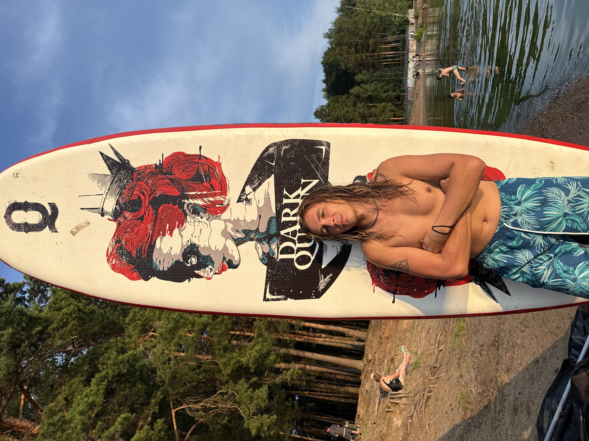 A man with long, blonde hair stands shirtless in front of a paddleboard. The paddleboard features a graphic of a woman with red hair and a crown, along with the words "DARK QUEEN." The man is wearing blue patterned swim trunks and has his arms crossed. He is positioned on a sandy shore next to a body of water. In the background, there are trees, people swimming in the water, and a few individuals sitting on the shore. The sky is blue with some clouds. The scene appears to be a recreational area, possibly a lake or beach, on a sunny day.