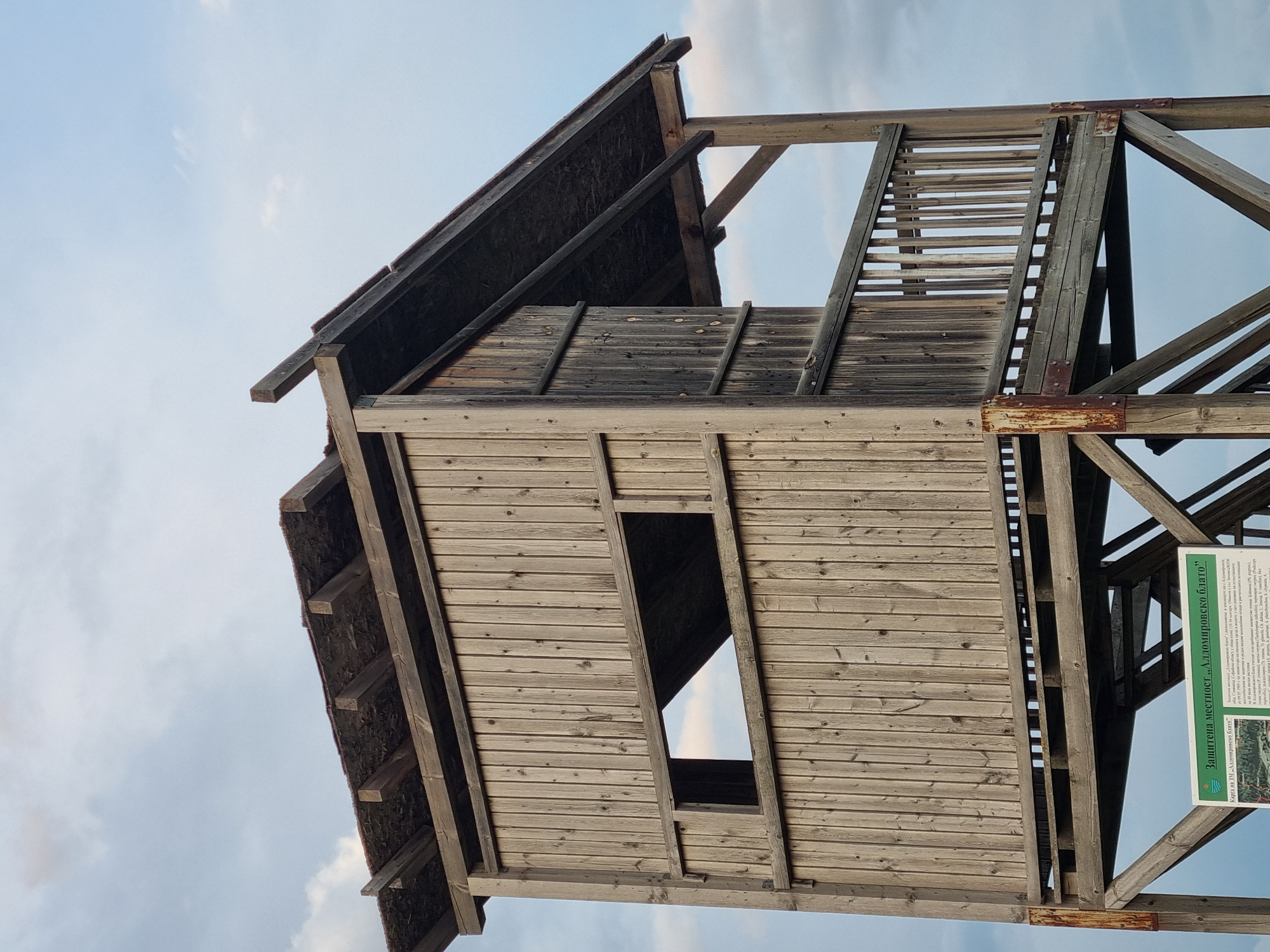 The image captures an upward view of a wooden observation tower, likely situated in or near the city of Aldomirovtsi, Bulgaria, based on the signage visible in the lower right corner which mentions "Защитена местност „Алдомировско блато"", which translates to "Protected area 'Aldomirovsko blato'". The tower features a multi-tiered construction with a sloped roof. The wooden planks exhibit signs of weathering and aging. The tower's structure includes visible support beams and a ladder-like section with horizontal rungs. The sky provides a backdrop, with a mix of blue and cloudy patches. The sign is green with white text and includes a map.