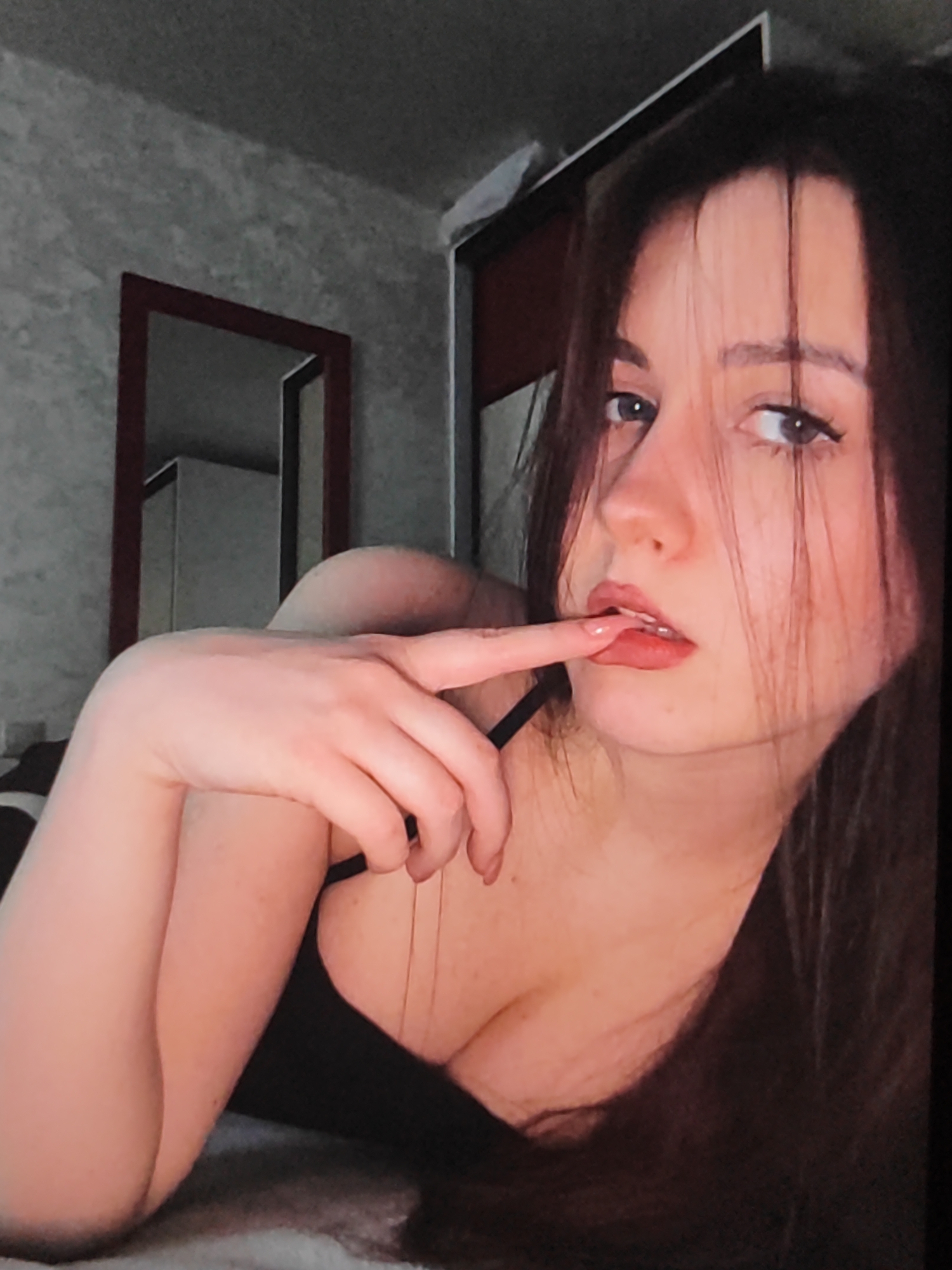 The image depicts a young woman with dark hair, lying on a bed with her head propped up. She is wearing a black top and has her finger gently touching her lips. Her gaze is directed towards the viewer. In the background, a mirror with a dark frame is visible, reflecting a wardrobe. The walls have a textured, grey wallpaper. The overall setting appears to be a bedroom, likely within a residential apartment in Fergana City, Uzbekistan, given the request.