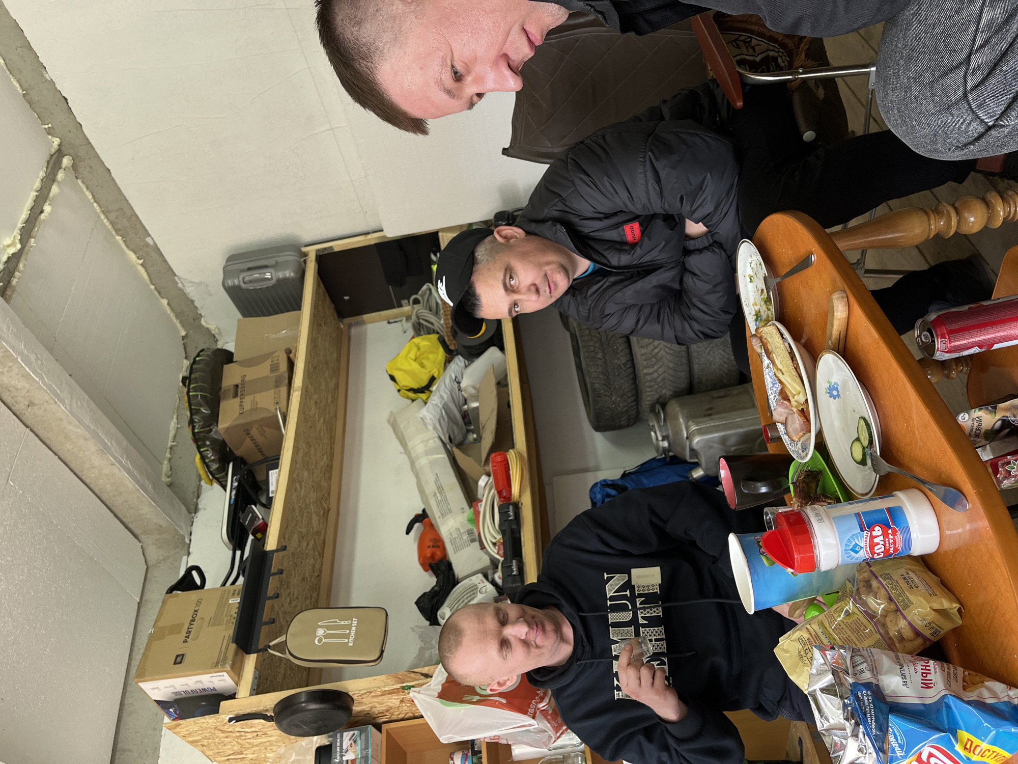 The scene depicts three men sitting at a table laden with food and condiments inside what appears to be a storage room or garage. The room has exposed insulation on the walls and shelving units filled with boxes, tools, tires, and miscellaneous items.
The table is made of light-colored wood and is crowded with opened bags of chips, a salt shaker labeled "EXTRA SALT", plates with food remnants, and a fork. One man is seated at the bottom of the frame, wearing a black hoodie with text, holding a glass, and looking directly at the camera. Another man, dressed in a black jacket, sits near the middle, with his arms crossed, also looking at the camera. A third man, wearing grey, leans into the frame from the top right corner.
Boxes labeled "PARTYBOX 100" and "KITCHEN SET" are visible on the shelves. The overall atmosphere is casual and possibly festive. The OCR suggests the image was taken in or relates to Chaikovsky, Russia, due to the Russian text on the products and packaging.