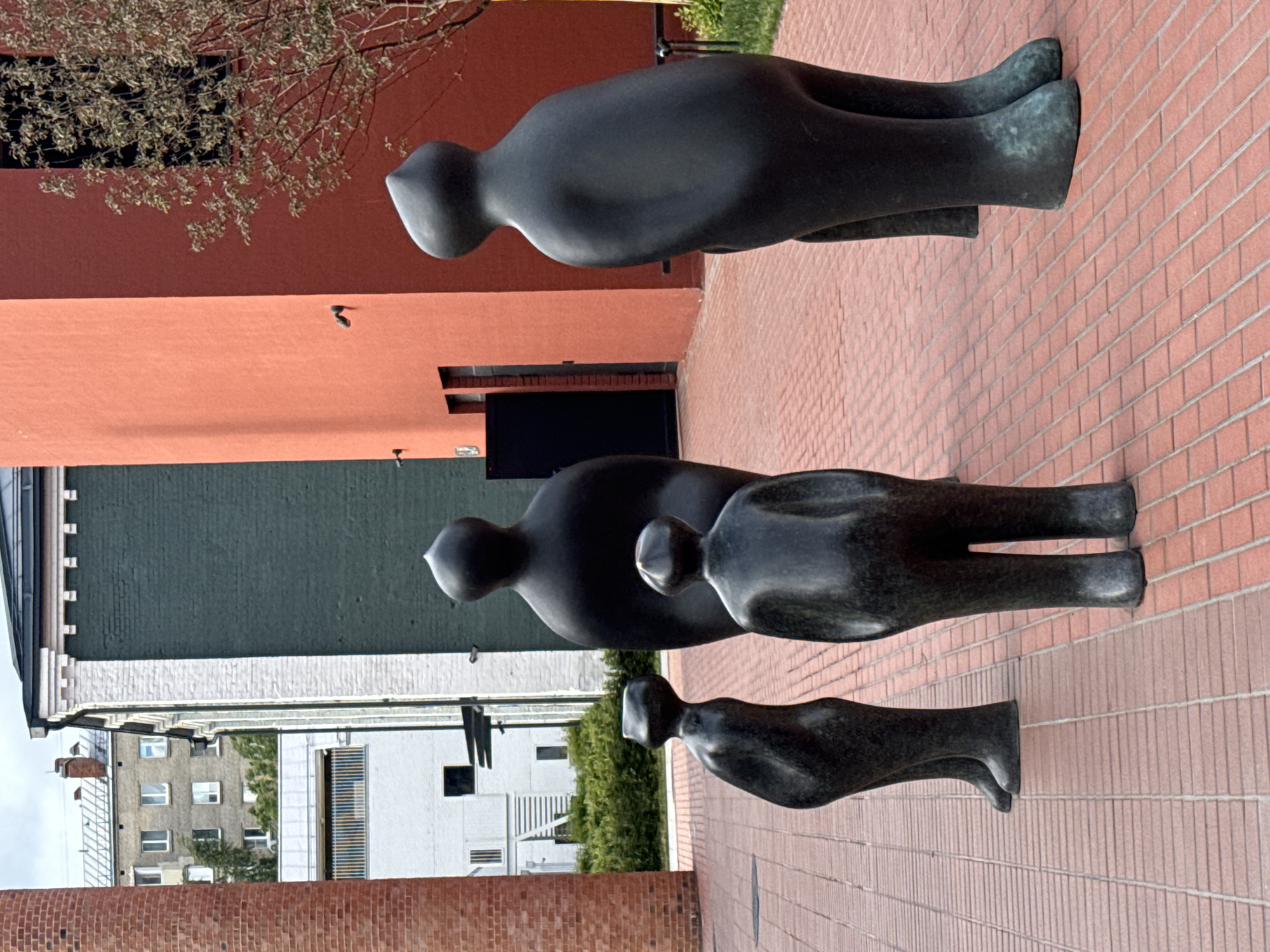 The image shows a sculpture in Moscow, Russia. The sculpture depicts three stylized human figures, a larger one followed by a smaller one, and then another one following the smaller one. The figures are black or dark bronze, and are arranged in a line along a brick walkway. Behind the sculptures, there is a building with sections of different colors (red, green, and white), and also distant buildings. A tree's branches are visible above the sculptures. The orientation of the image suggests it has been rotated.