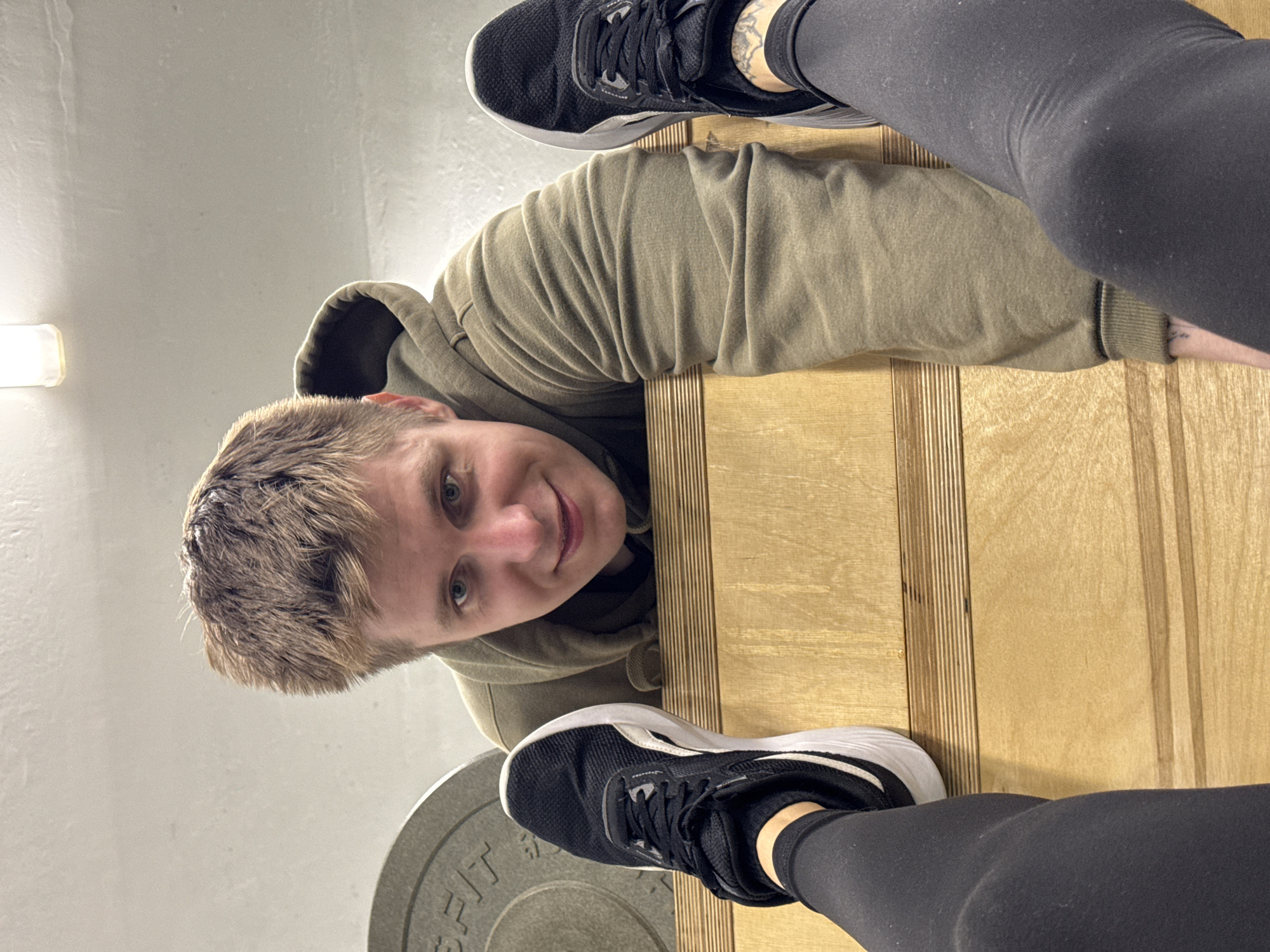 An indoor, low-angle image, possibly taken in Chaikovsky, Russia, shows a young person with short, light brown hair and blue eyes looking directly at the camera with a slight, friendly smile. They are wearing an olive green hooded sweatshirt and appear to be lying or leaning on a light-colored wooden surface, which features distinct vertical planks.
The foreground is dominated by the feet and lower legs of another individual, likely the photographer. These legs are clad in black leggings, and the feet wear black athletic shoes with white soles. One foot is visible in the upper right, and the other in the lower left, partially resting on a grey circular weight plate. The word "FIT" is partially visible on the weight plate. The background features a plain, light-colored wall, with a bright, rectangular light fixture visible on the left side. The overall setting suggests an indoor fitness environment, such as a gym.