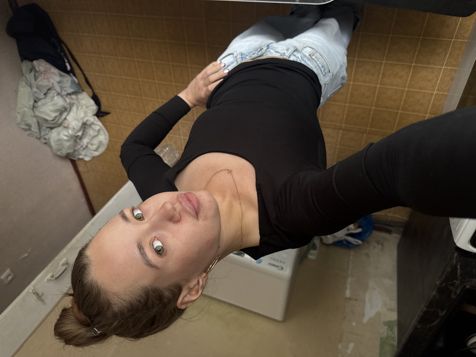 A woman is taking a selfie in a room that appears to be a bathroom or laundry room. She is wearing a black long-sleeved shirt and light-wash jeans. Her hair is pulled up in a bun. She is wearing large hoop earrings and a thin necklace. The room has tiled flooring and a pale yellow wall with peeling wallpaper in some areas. In the background, there is a white appliance, possibly a washing machine, and a pile of laundry on the floor next to a dark bag. The woman's pose suggests she is standing and looking down at her phone. The overall scene depicts a casual, indoor setting.