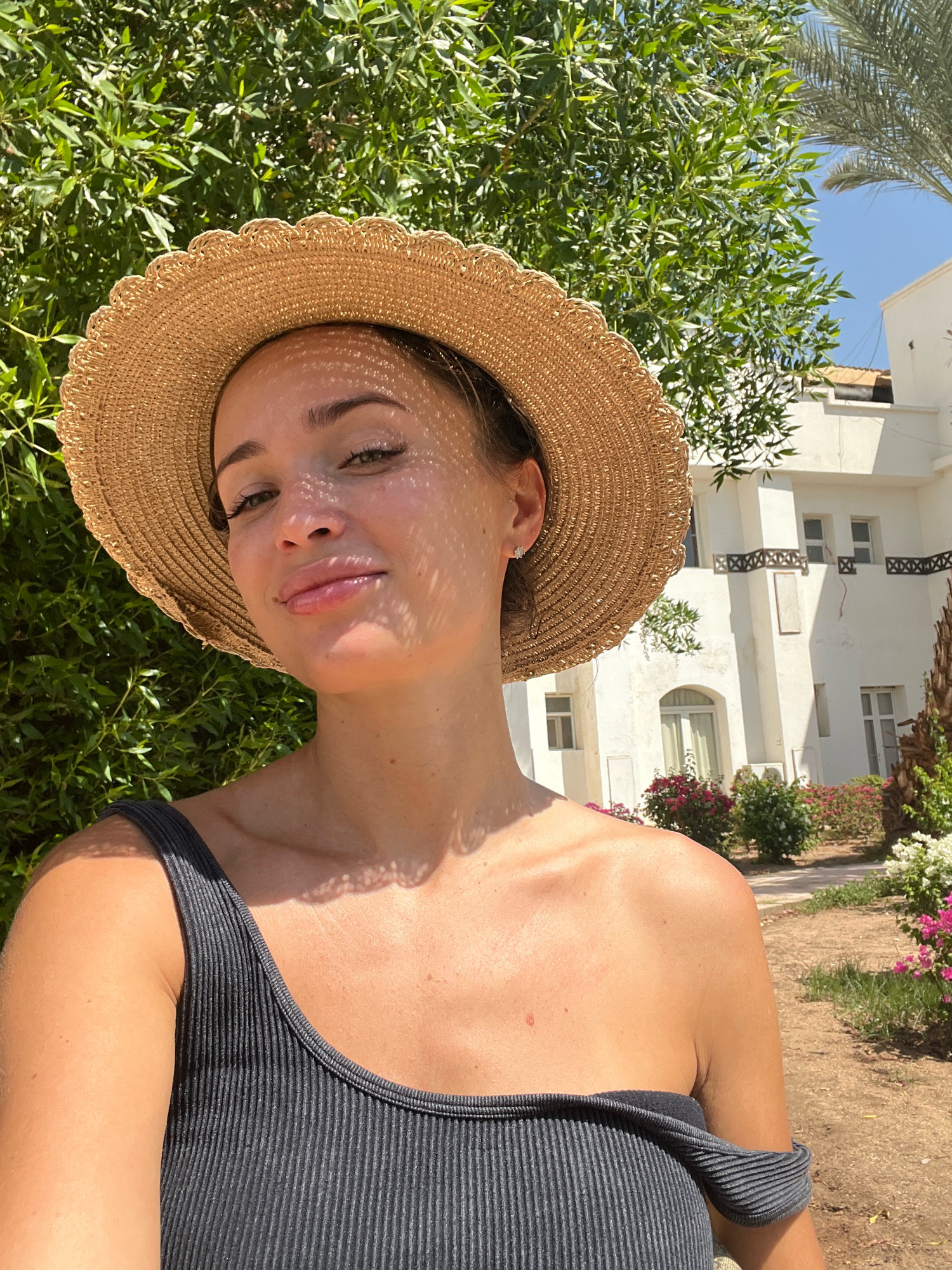 A woman wearing a straw hat and a dark grey ribbed swimsuit poses for a selfie outdoors. She has fair skin and is looking towards the camera with a slight smile. The background shows lush green foliage, a white building with balconies and windows, and a palm tree. The sunlight casts dappled shadows on her face and shoulders. The scene suggests a warm, sunny day in a resort or hotel environment, possibly in Sharm El Sheikh, Egypt, given the architecture and vegetation.