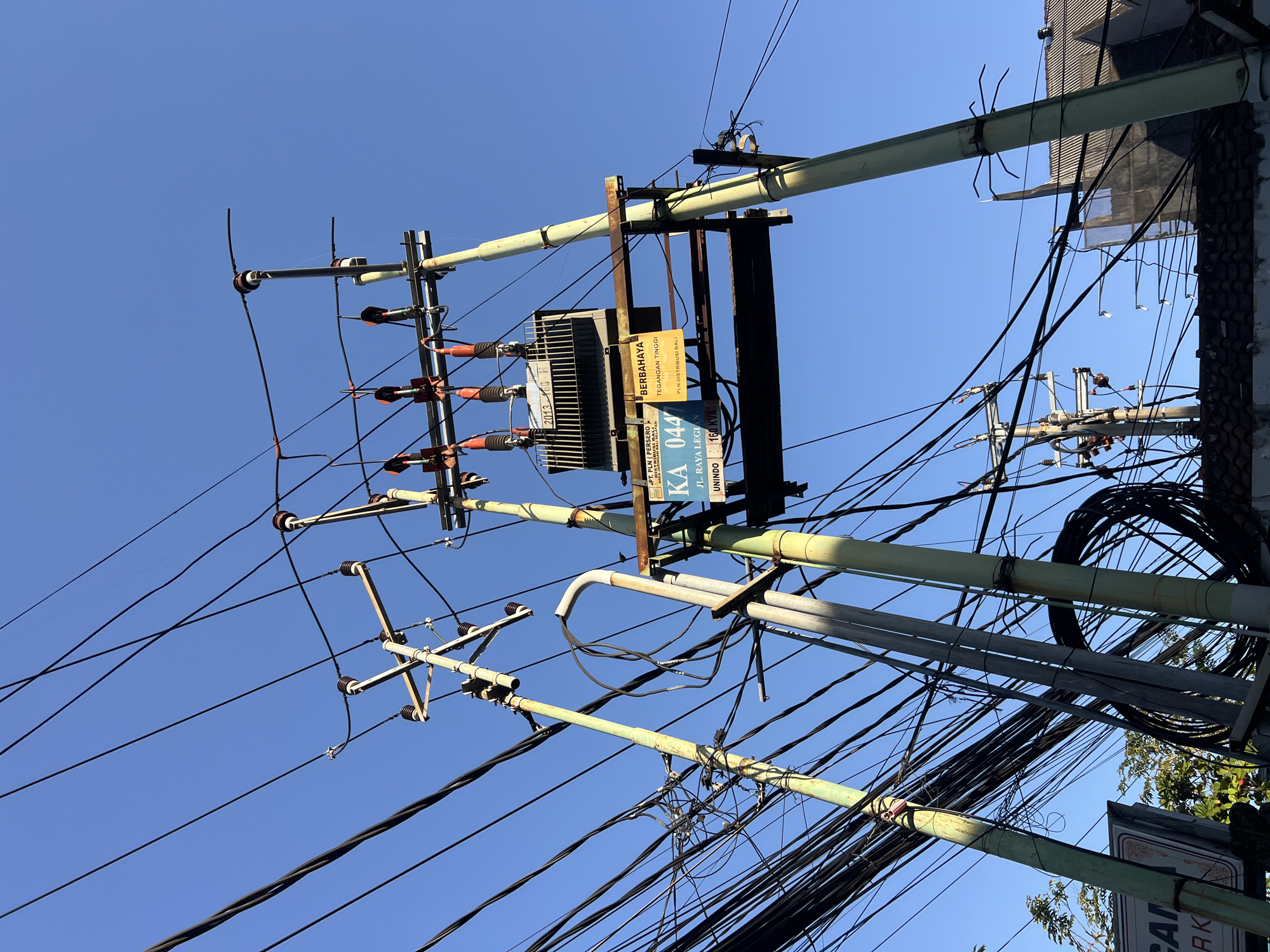 This is a ground-level, wide-angle, outdoor photograph taken on a clear day, likely in Kuta, Indonesia, showcasing a utility pole with a transformer and numerous power lines against a bright blue sky. The pole itself appears to be made of metal and is painted a pale green, showing signs of rust and wear. A large electrical transformer is mounted on the pole, connected to a dense network of black electrical wires that crisscross the frame, creating a visually complex and somewhat chaotic arrangement.
Two yellow signs are attached to the pole. One larger sign, partially obscured, has the text "PT. PLN (PERSERO)" followed by what appears to be Indonesian text. Below this, a smaller sign reads "KA 044" and further text that includes "JL. RAYA LEGIAN" and "UNINDO 160 KVA". A second, smaller yellow sign to the right reads "BERBAHAYA TEGANGAN TINGGI" which translates to "DANGER HIGH VOLTAGE".
The scene is dominated by the utility infrastructure, with no people visible. The background shows a sliver of rooftops of buildings with tiled roofs on the right side of the image. The overall impression is one of densely populated urban infrastructure.