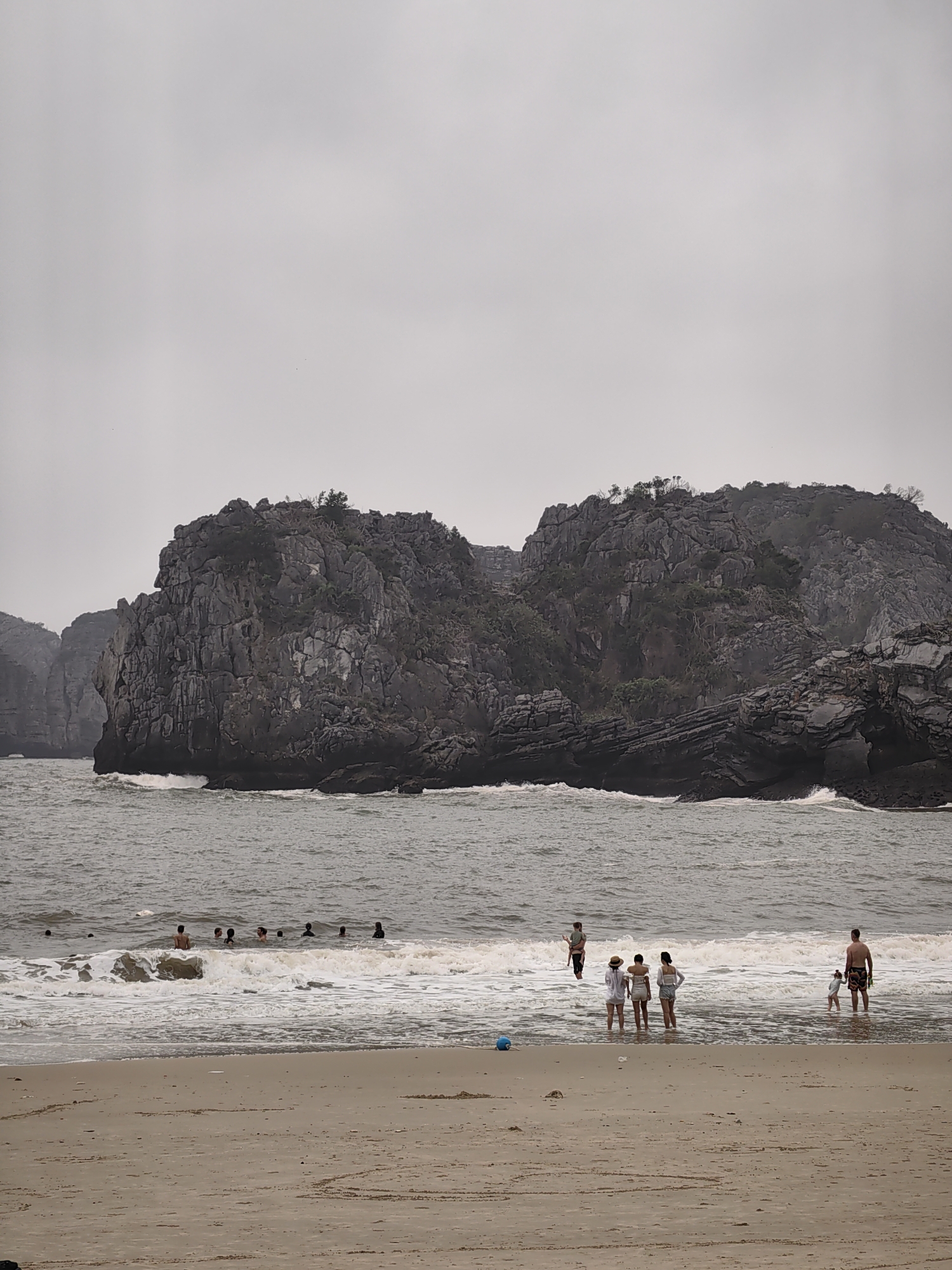 The image shows a beach scene in Hải Phòng, Vietnam, on an overcast day. A large, rocky island or headland rises from the sea in the background. In the water near the shore, a group of people are swimming or wading. Closer to the beach, another small group stands at the water's edge. A blue ball rests on the sand near them. A man and a child are further out in the water. The sand is wet near the water line, and a large heart is drawn into the sand near the bottom of the image.