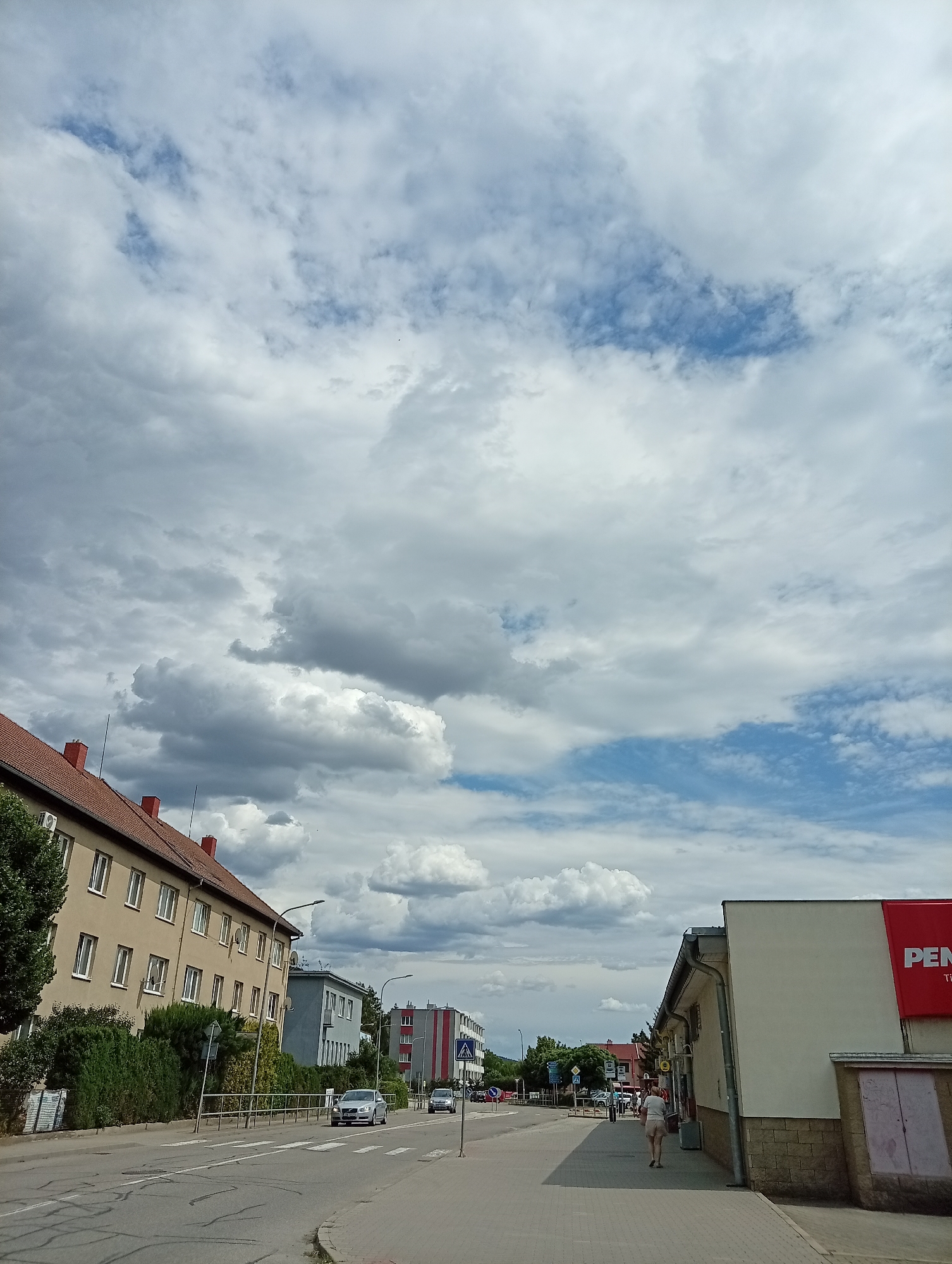This is a vertical, eye-level, outdoor photograph taken on a street in Tišnov, Czechia. The sky is filled with a mix of white and gray clouds, with patches of blue visible. On the left side of the street, a multi-story building with a red tile roof and beige siding runs along the roadway. The building has many windows and is bordered by dense green bushes. Across the street, several cars are driving, and a crosswalk is painted on the asphalt. Further down the street, more buildings can be seen, including one with a red and white striped facade. On the right side of the image, a building with a white facade and a large red sign that partially reads "PEN" is visible. A person wearing a white shirt and light-colored shorts is walking away from the camera on the sidewalk in front of this building. The street is paved with asphalt and the sidewalk is made of interlocking pavers. The overall impression is of a typical European town street on a cloudy day.