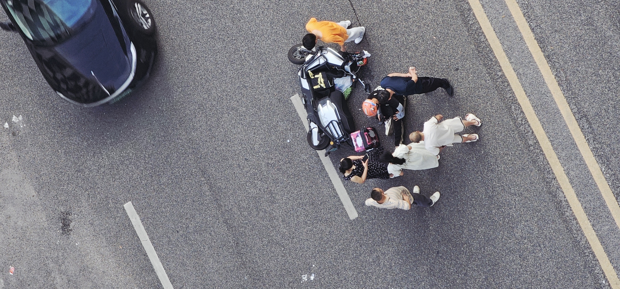 The image, captured from an aerial perspective, depicts an apparent vehicular accident scene in Jiulongpo District, China. A black car is positioned at the upper left corner of the frame, while a toppled white motorcycle lies on its side, surrounded by several individuals. One person is dressed in an orange outfit, and another wears an orange helmet, suggesting they might have been riding the motorcycle. One man is kneeling and leaning over the person in the helmet. Several people are lying or kneeling near the motorcycle, while another man stands upright at the edge of the group. A woman sitting on the ground is holding a cell phone. The accident occurred on a gray asphalt road featuring white painted lane markers on one side and double yellow lines on the other. The immediate environment is the road surface, with no visible buildings or other landscape features in the background.