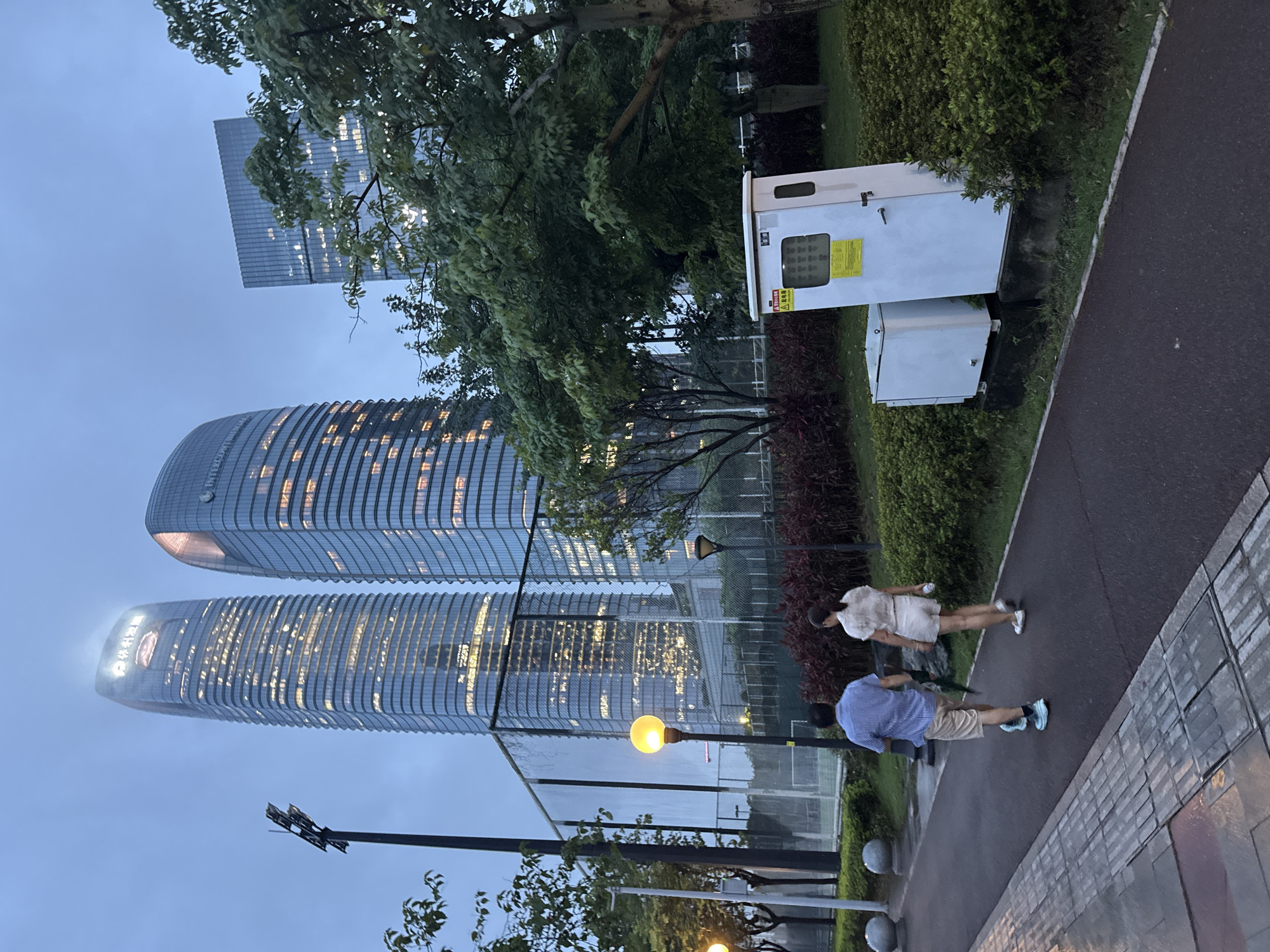 In Haizhu District, China, two individuals, a male and a female, walk away from the camera on a paved pathway beside a lush green area with trees and bushes. The male, wearing a checkered blue shirt and light-colored shorts, is walking slightly ahead of the female, who is wearing a light-colored top and shorts. They appear to be strolling in the early evening or twilight, as indicated by the dim natural light and the illuminated windows of several modern skyscrapers in the background.
The skyscrapers, characterized by their glass facades and illuminated interiors, dominate the skyline. One building in the foreground on the left displays a prominent logo at its apex. To the left of the pathway, a chain-link fence encloses what appears to be a sports court, possibly a tennis court, with a light pole nearby. On the right side of the pathway, a white utility box with warning labels is situated among the greenery. The ground is damp, suggesting recent rain or dew. The overall atmosphere is urban and serene, with the juxtaposition of natural elements and towering architecture.
